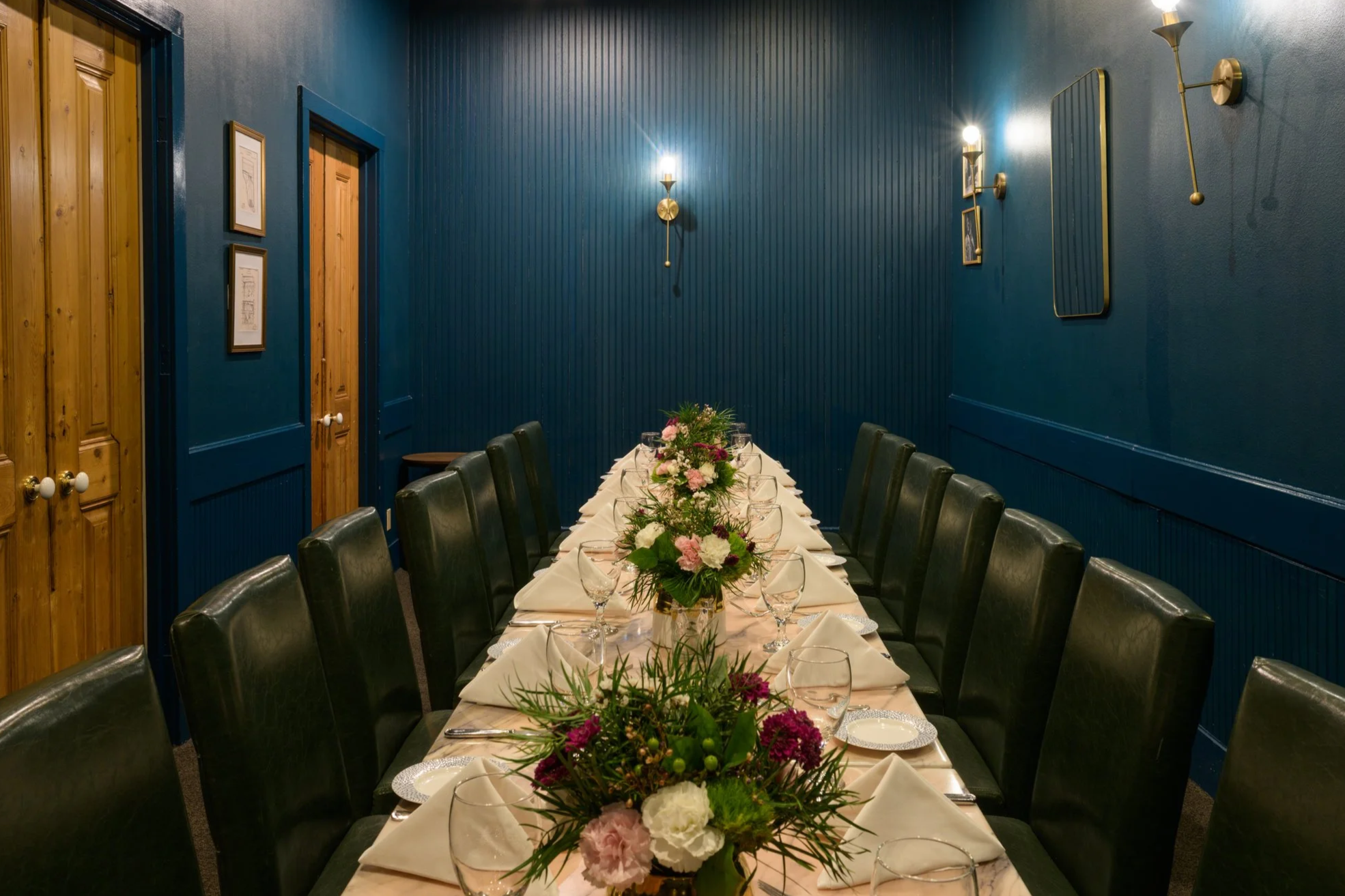 Elegant dining room with a long table set for a formal meal. The room has a dark blue wall with decorative sconces, mirrors, and framed art. The table is adorned with white napkins, glassware, and floral centerpieces. A flat-screen TV is mounted on the wall.
