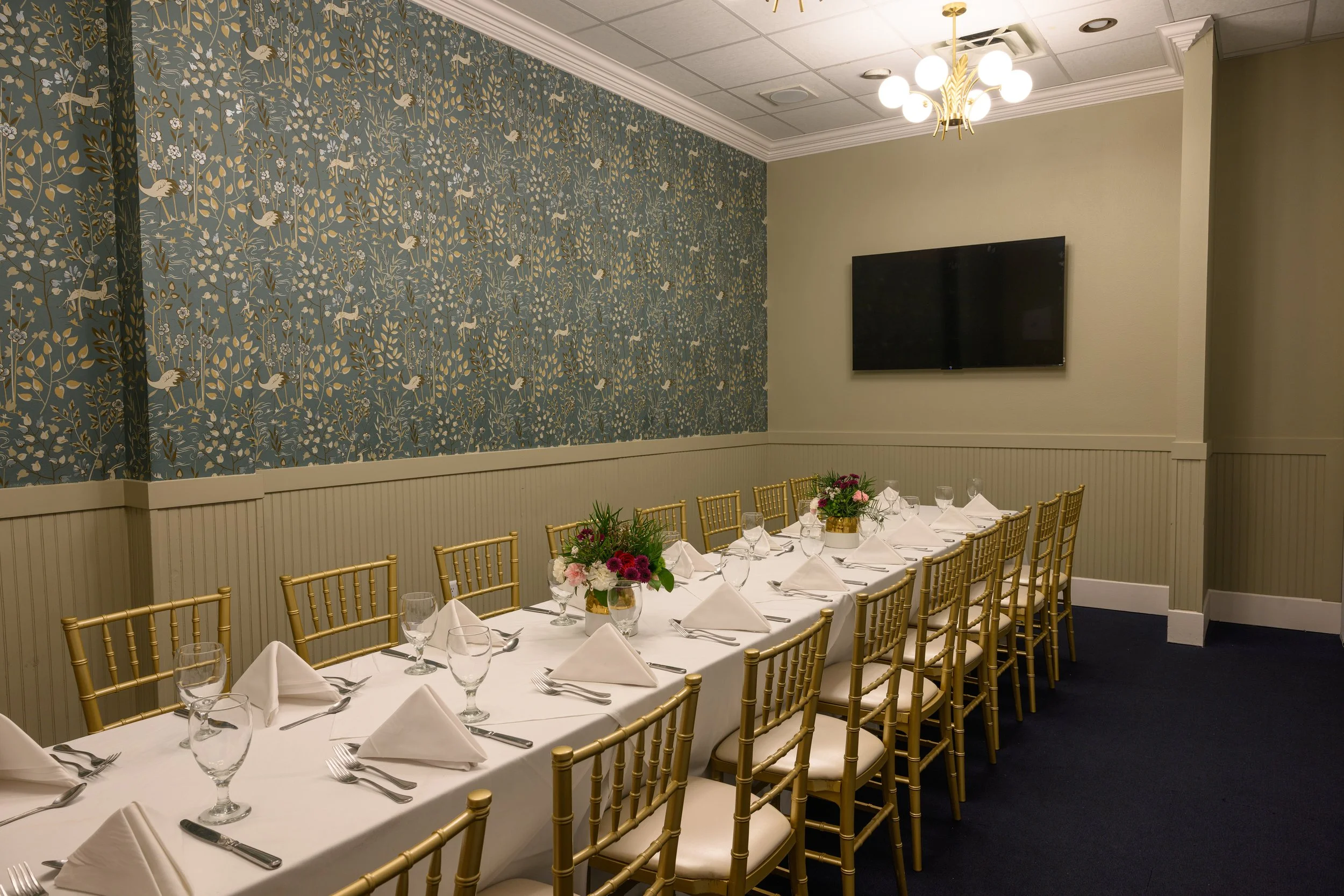 A long dining table in a room with floral wallpaper, set for a meal with chairs on both sides, a white tablecloth, and flower centerpieces. There is a flat-screen TV on the wall and a modern chandelier overhead.