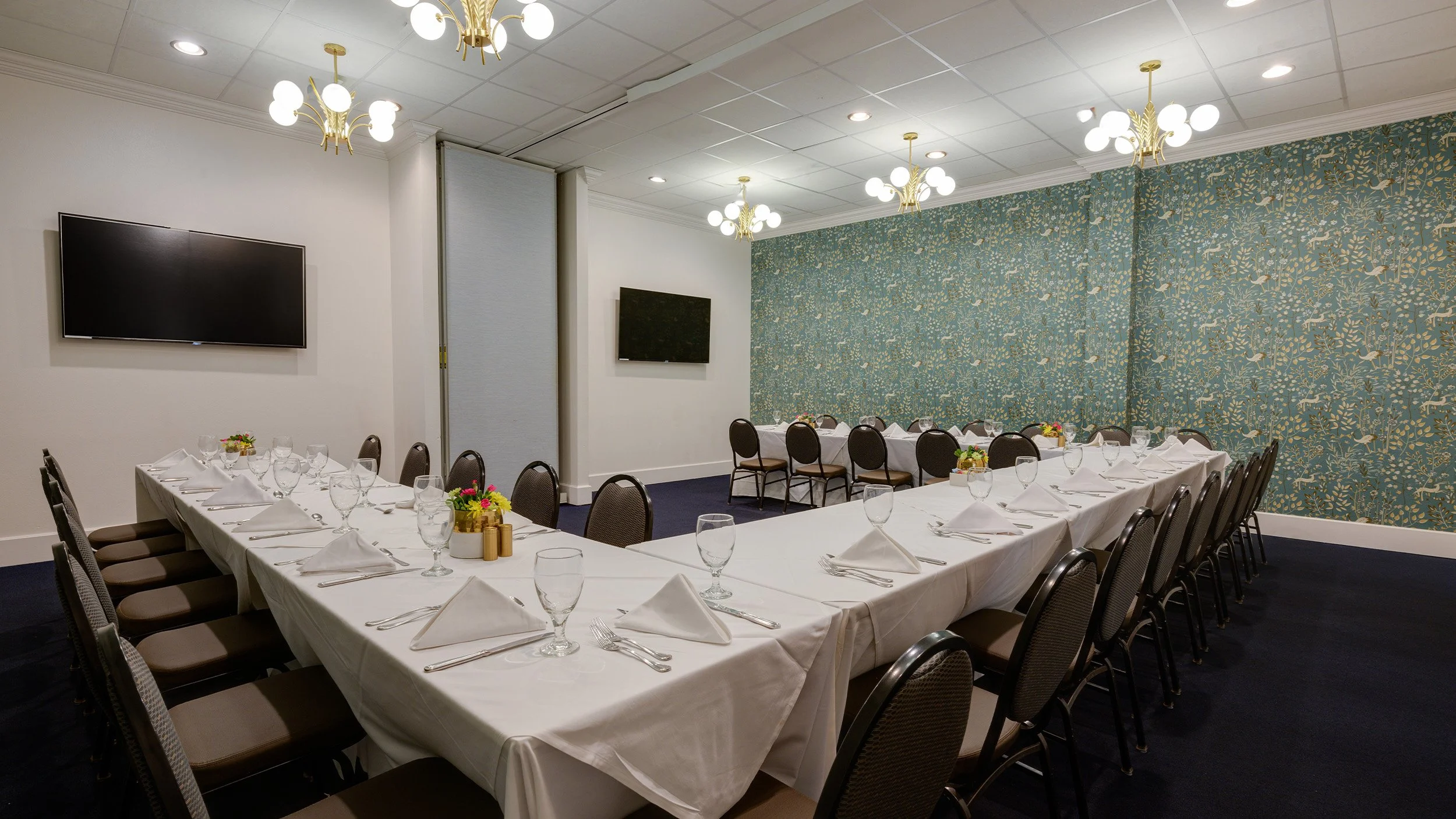 Conference room with long table and chairs, set for a formal meeting or banquet, featuring white tablecloths, glassware, and floral centerpieces, with two televisions on the walls.
