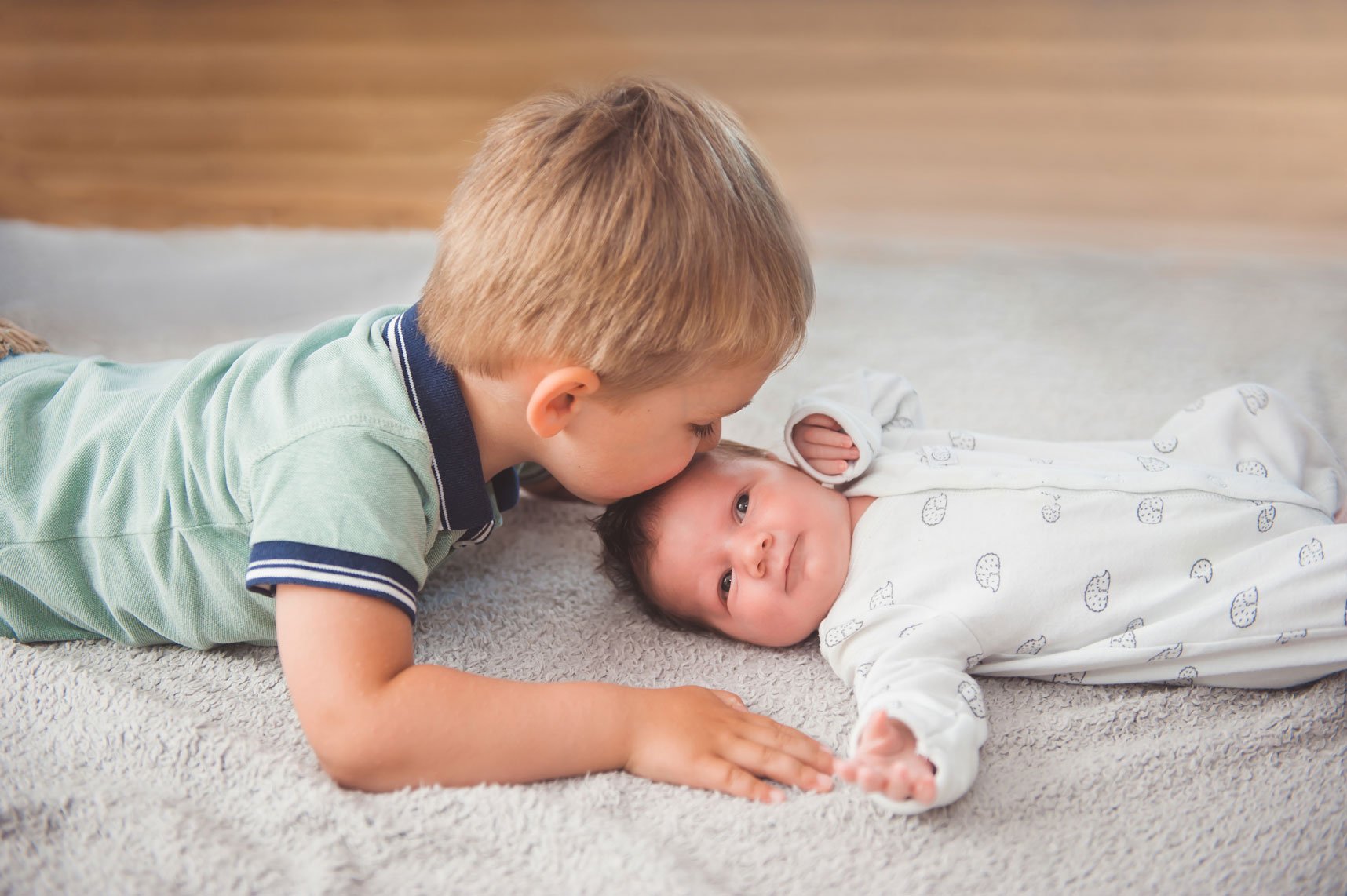 Ein kleiner Junge küsst seine Schwester auf die Stirn. Das Baby liegt auf einem Teppich und trägt weiße Kleidung mit einem Muster. Sie schauen sich zärtlich an.