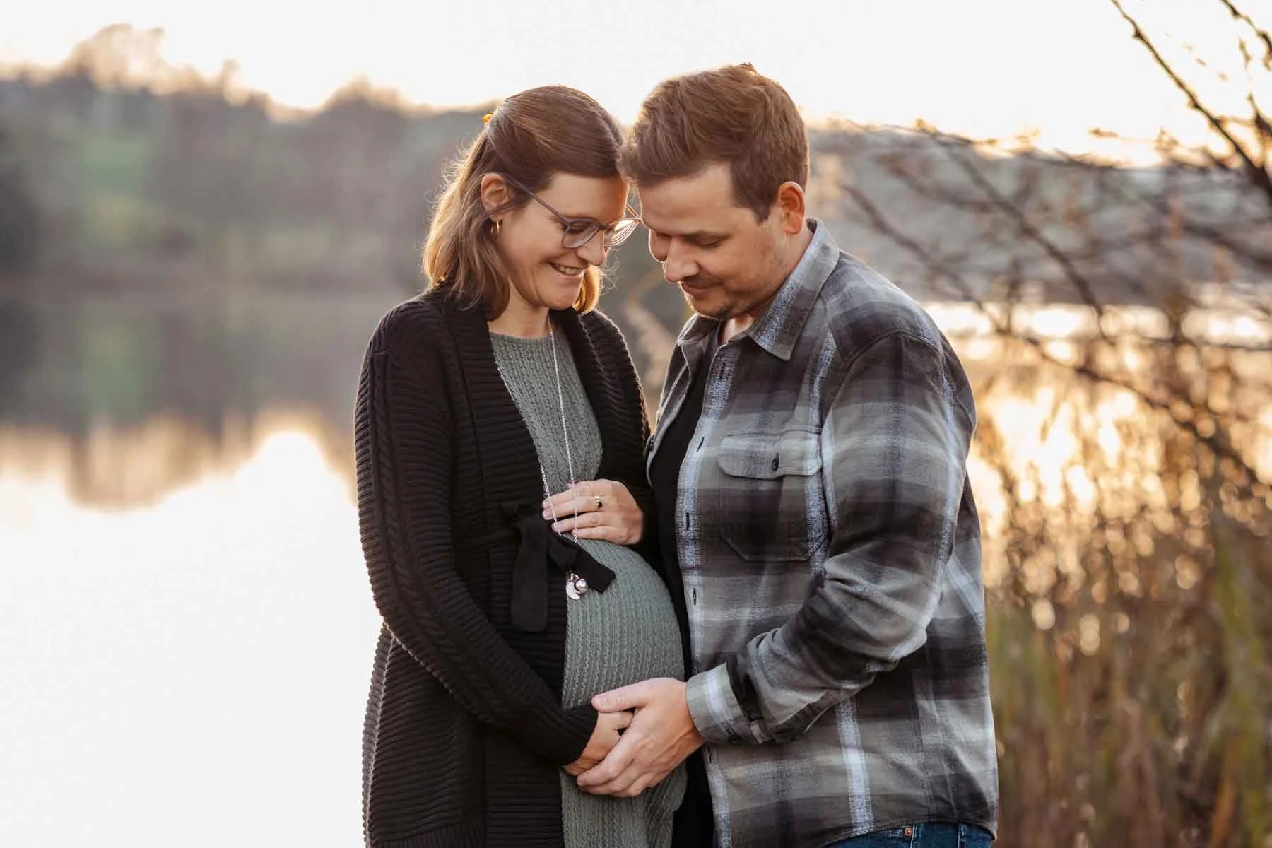 Ein Paar mit Babybauch steht im Freien an einem See bei Sonnenuntergang und hält Händchen.
