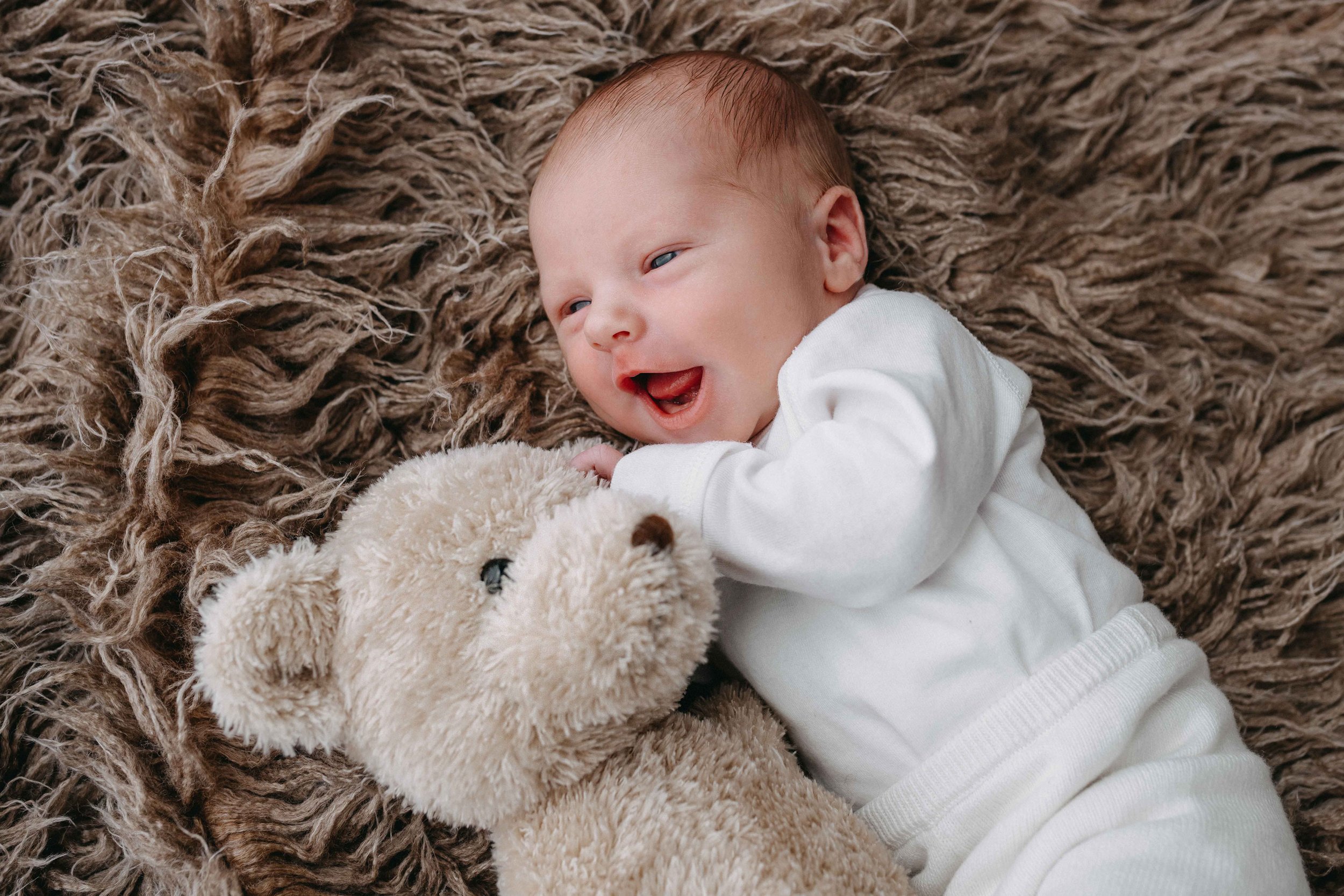 Lächelndes Baby in weissem Strampler auf kuscheligem braunem Teppich, mit einem Plüschteddy.