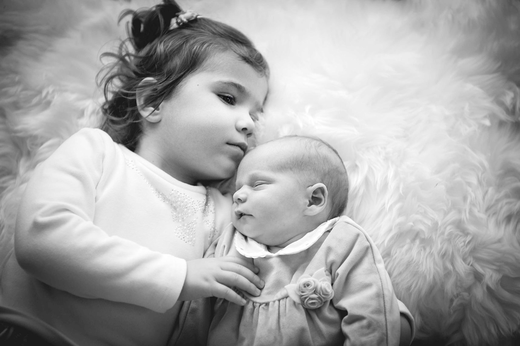 Ein kleines Mädchen liegt neben einem schlafenden Baby auf einem flauschigen Hintergrund.