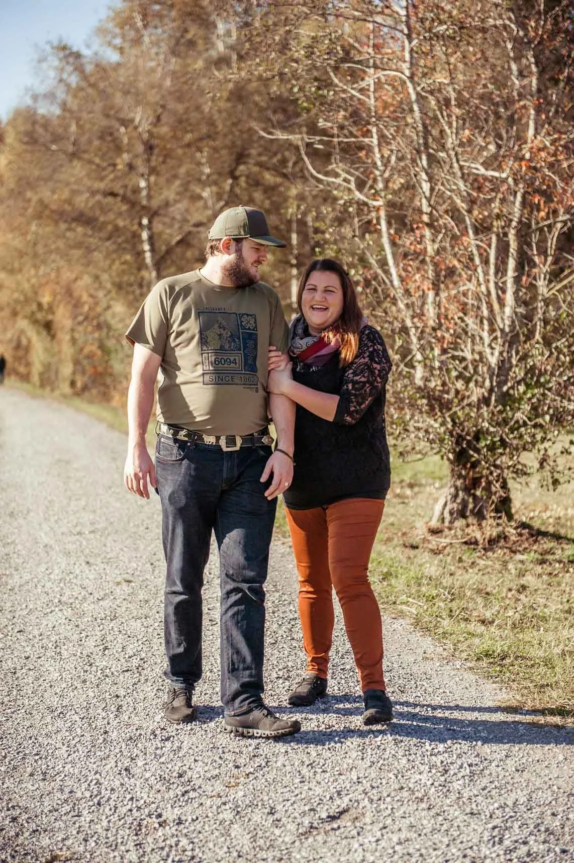 Ein lachendes Paar spaziert auf einem Wanderweg im Herbst, umgeben von Bäumen mit goldbraunen Blättern.