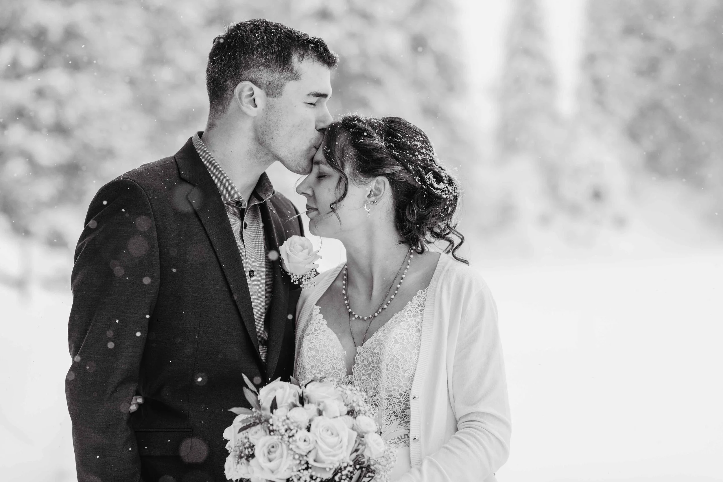 Ein Brautpaar im Schnee, der Bräutigam küsst die Braut auf die Stirn, sie hält einen Blumenstrauss.