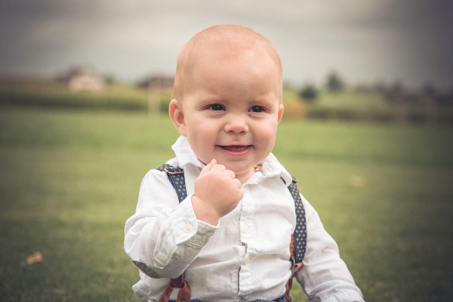 Ein lächelndes Baby mit weissem Hemd und Hosenträgern auf einem Grasfeld.