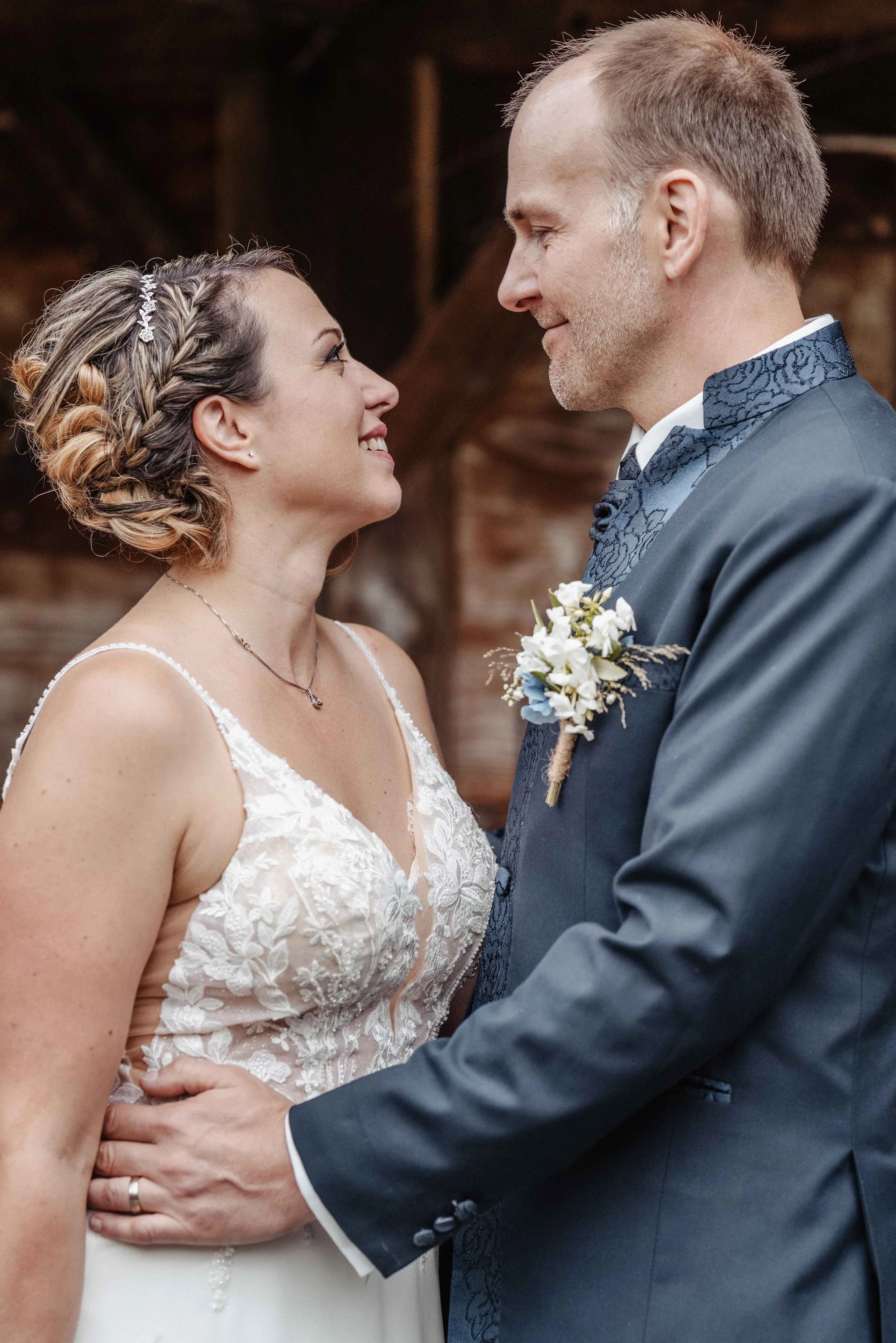 Ein verliebtes Brautpaar blickt sich in die Augen, die Braut trägt ein weisses Spitzenkleid, der Bräutigam einen dunkelblauen Anzug.