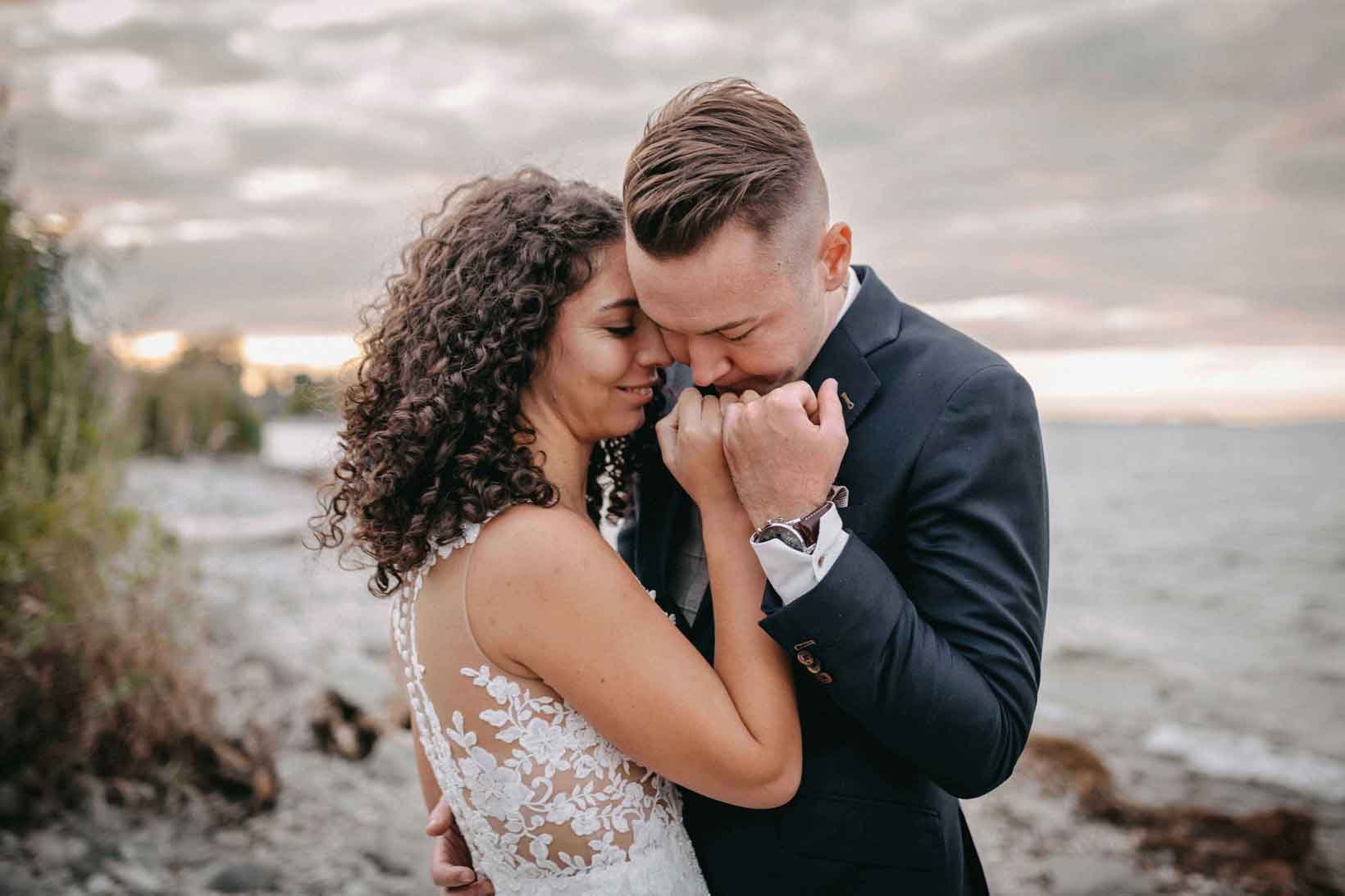 Ein glückliches Paar, eine Frau mit lockigem schwarzen Haar in einem weissen, verzierten Kleid und ein Mann in einem schwarzen Anzug, stehen am Strand bei Sonnenuntergang, halten Händchen und liegen sich an den Lippen.