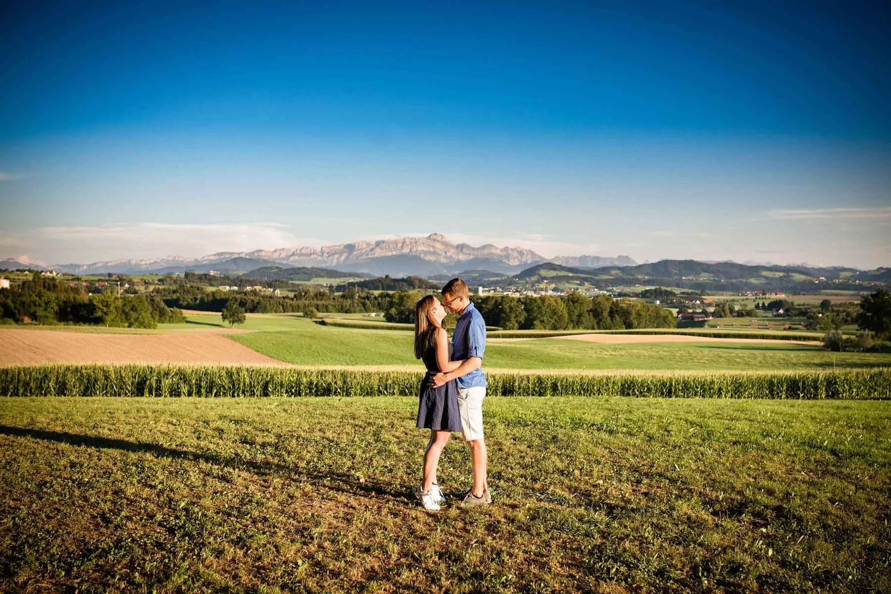 Paar steht in einer grünen Felderlandschaft mit Bergen im Hintergrund, umarmt sich und schaut sich an.
