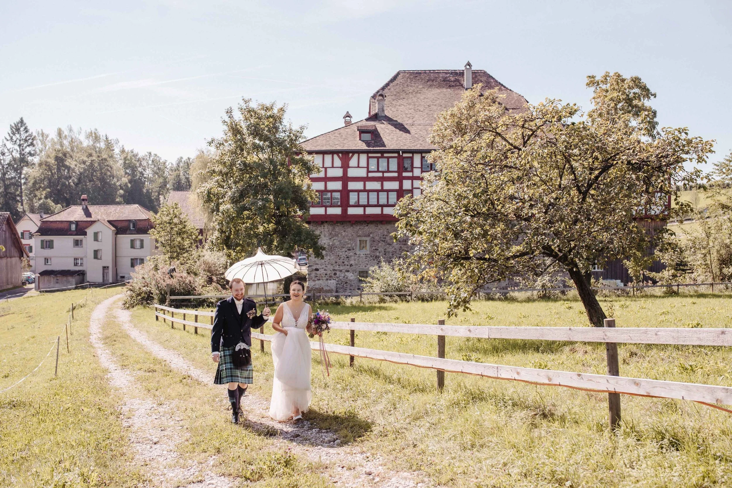 Hochzeitspaar, das auf einem Weg in der Natur spazieren geht, mit einem weißen Regenschirm und einem Blumenstrauss, umgeben von Bäumen und einem historischen Gebäude im Hintergrund.