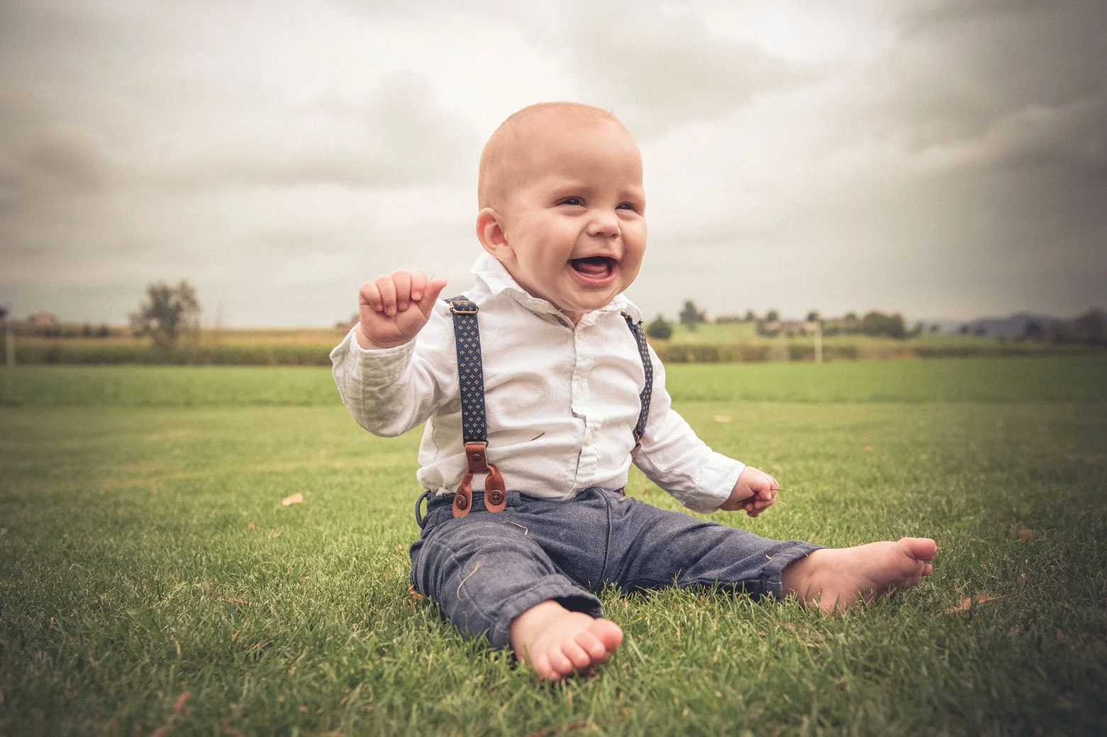 Lächelnder Babyjunge sitzt barfuss auf einer grünen Wiese, bei bewölktem Himmel im Freien.