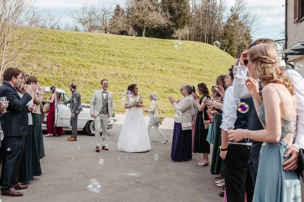 Ein Brautpaar wird von Freunden und Familie bei einer Hochzeit draussen gefeiert, Seifenblasen fliegen durch die Luft.