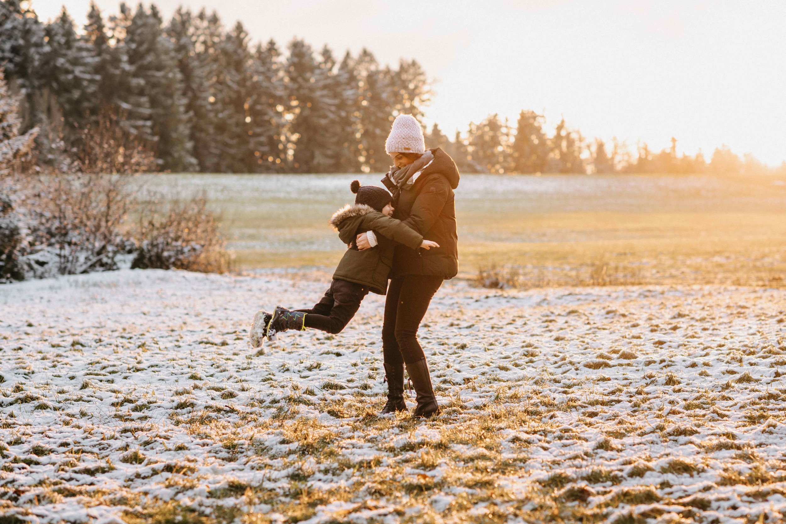Frau und Kind spielen auf schneebedecktem Feld mit Wald im Hintergrund. Sunset-Stimmung im Winter.