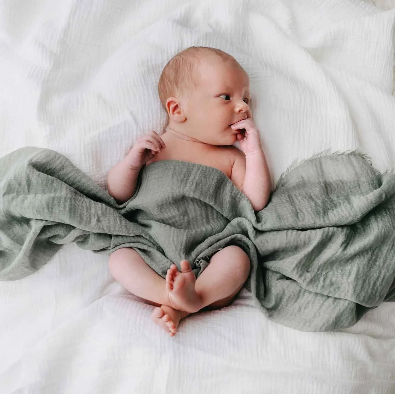 Ein neugeborenes Baby liegt auf einem weissen Bett, zur Hälfte in einer grüne Decke eingewickelt, mit nacktem Oberkörper. Das Baby schaut aufmerksam zur Seite.