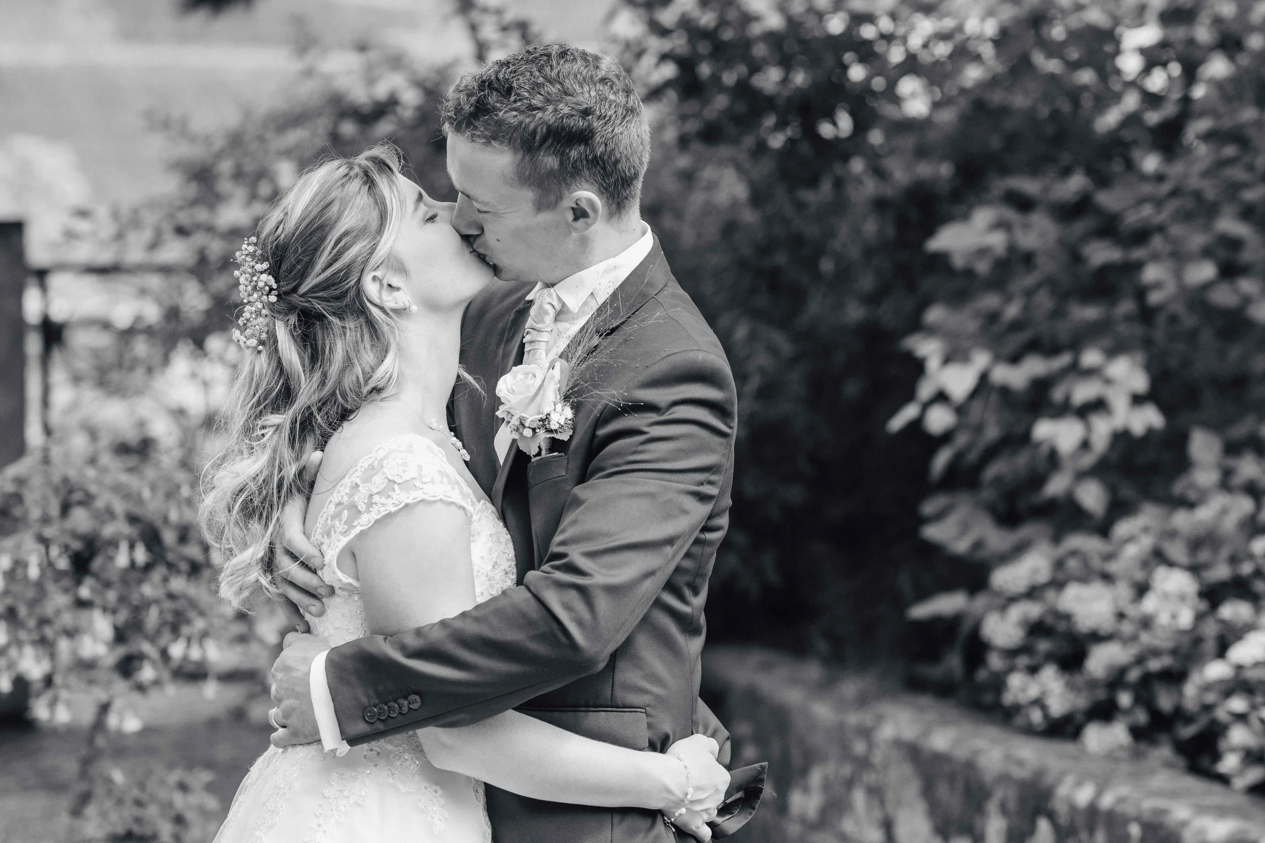 Braut und Bräutigam küssen sich im Freien, umgeben von Büschen und Blumen, schwarz-weisser Fotostil.