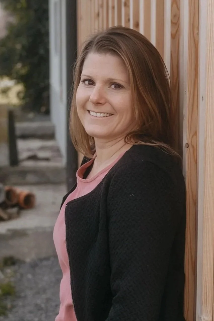 Janine Huber lacht und lehnt an einer Holzwand im Outdoor.