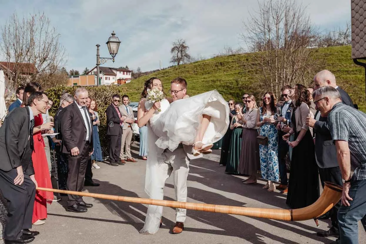 Braut wird von Bräutigam bei einer Hochzeit getragen, umgeben von Gästen, die zuschauen und fotografieren.