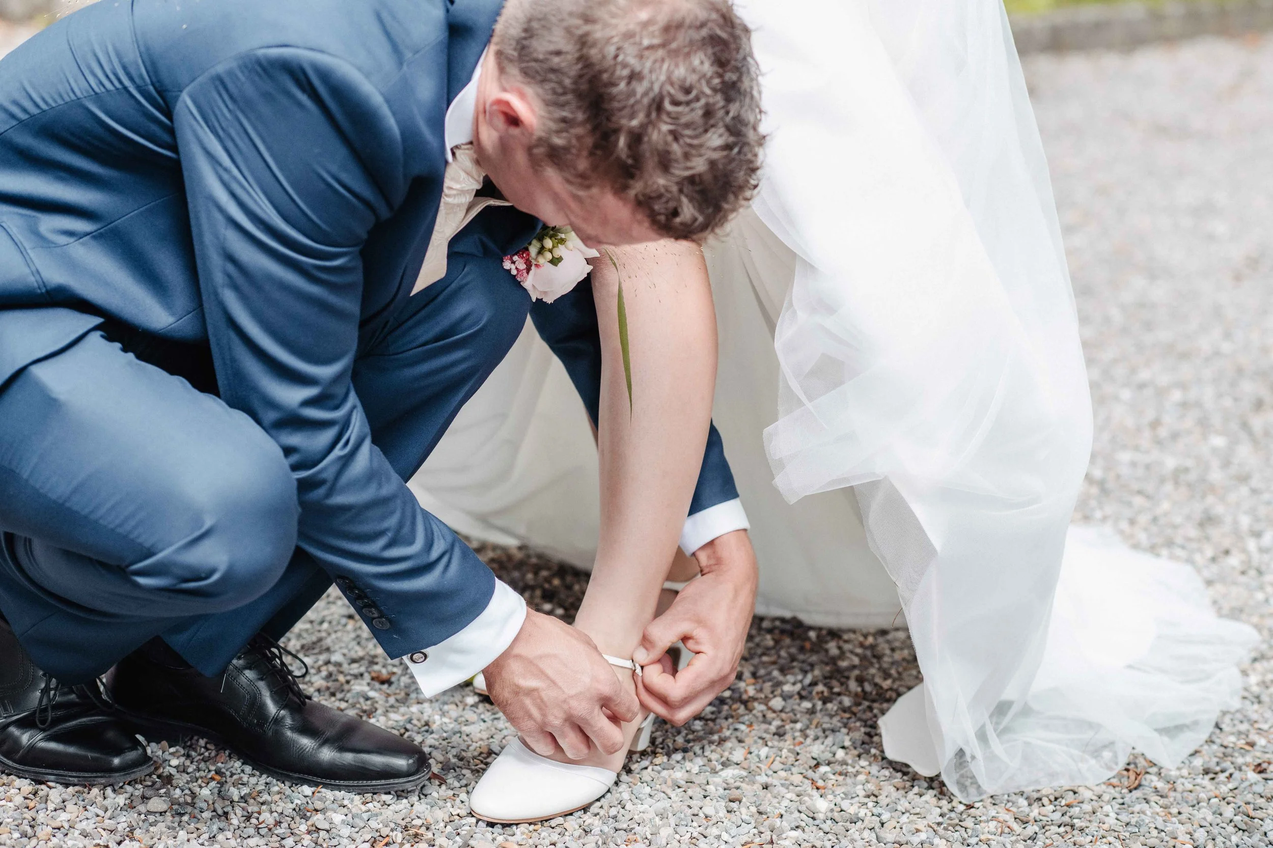 Ein Bräutigam hilft der Braut beim Anziehen der Schuhe auf kiesbedecktem Boden.