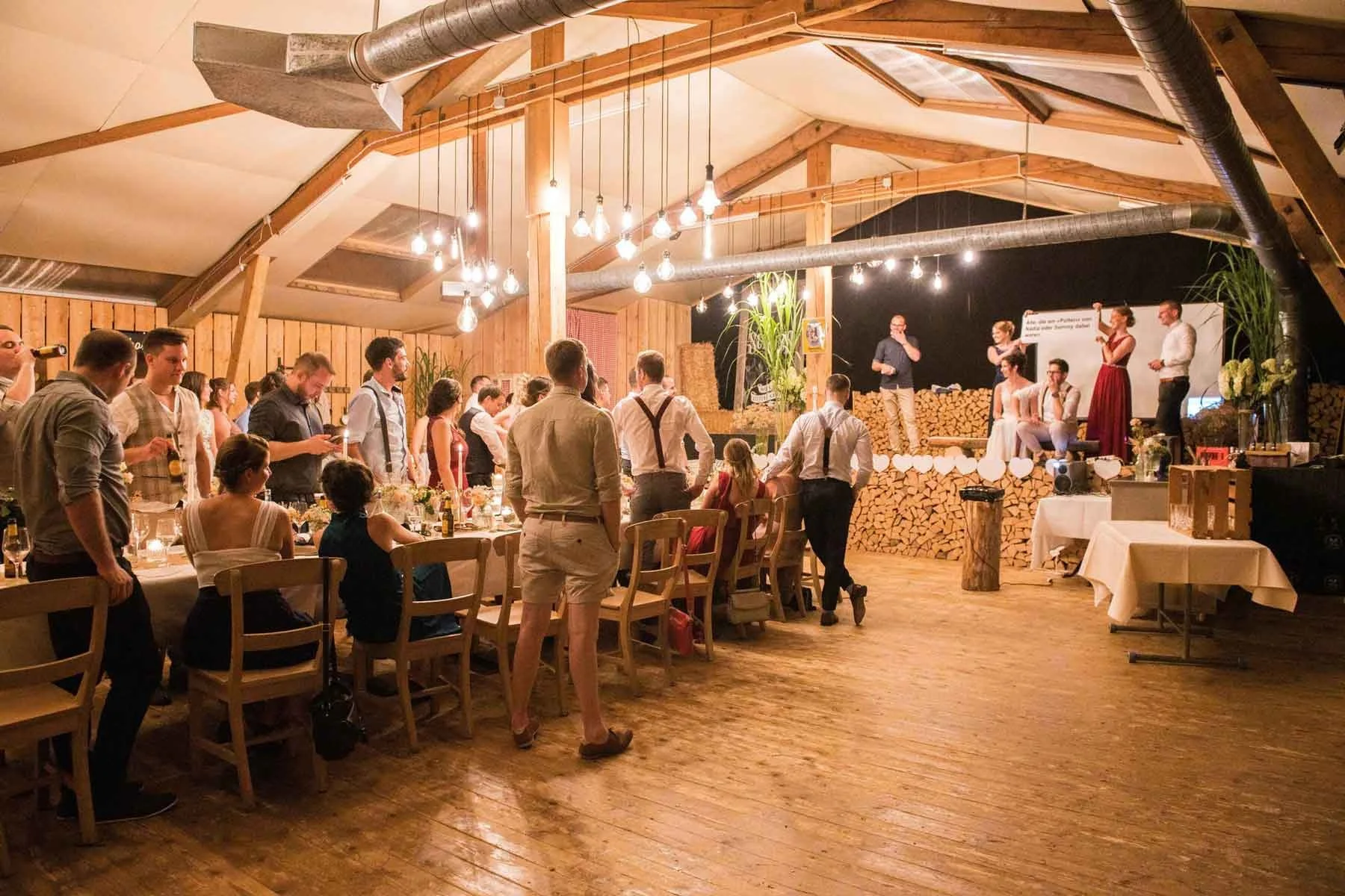 Ein Hochzeitsfest mit festlich dekorierten Tisch in einem rustikalen Veranstaltungsraum mit Holzdecke und beleuchteten Glühbirnen..