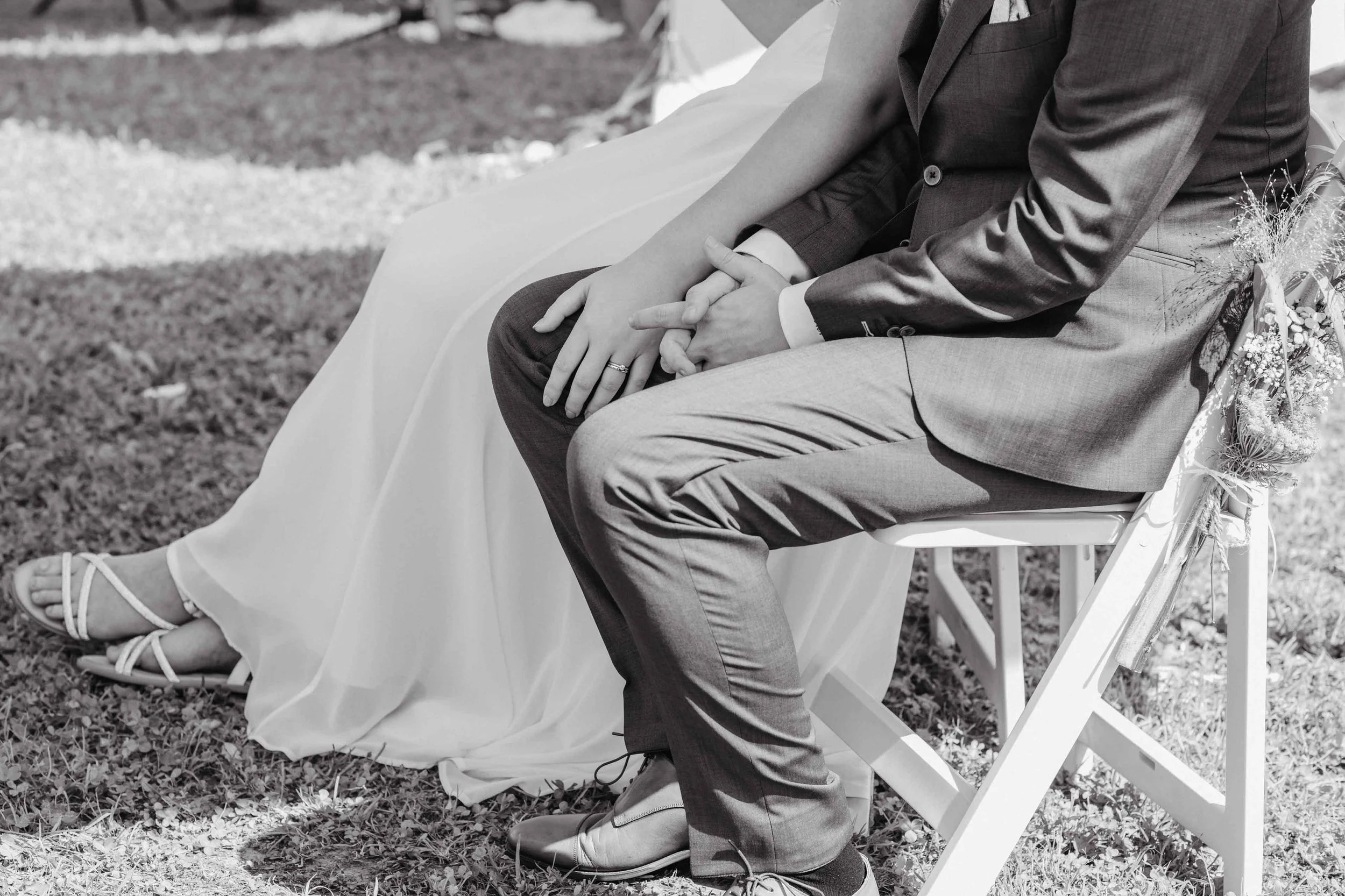 Ein Brautpaar sitzt auf einem Stuhl bei der freien Trauung, die Frau trägt ein weisses Hochzeitskleid und Sandalen, der Mann trägt einen dunklen Anzug. Sie halten sich an den Händen.