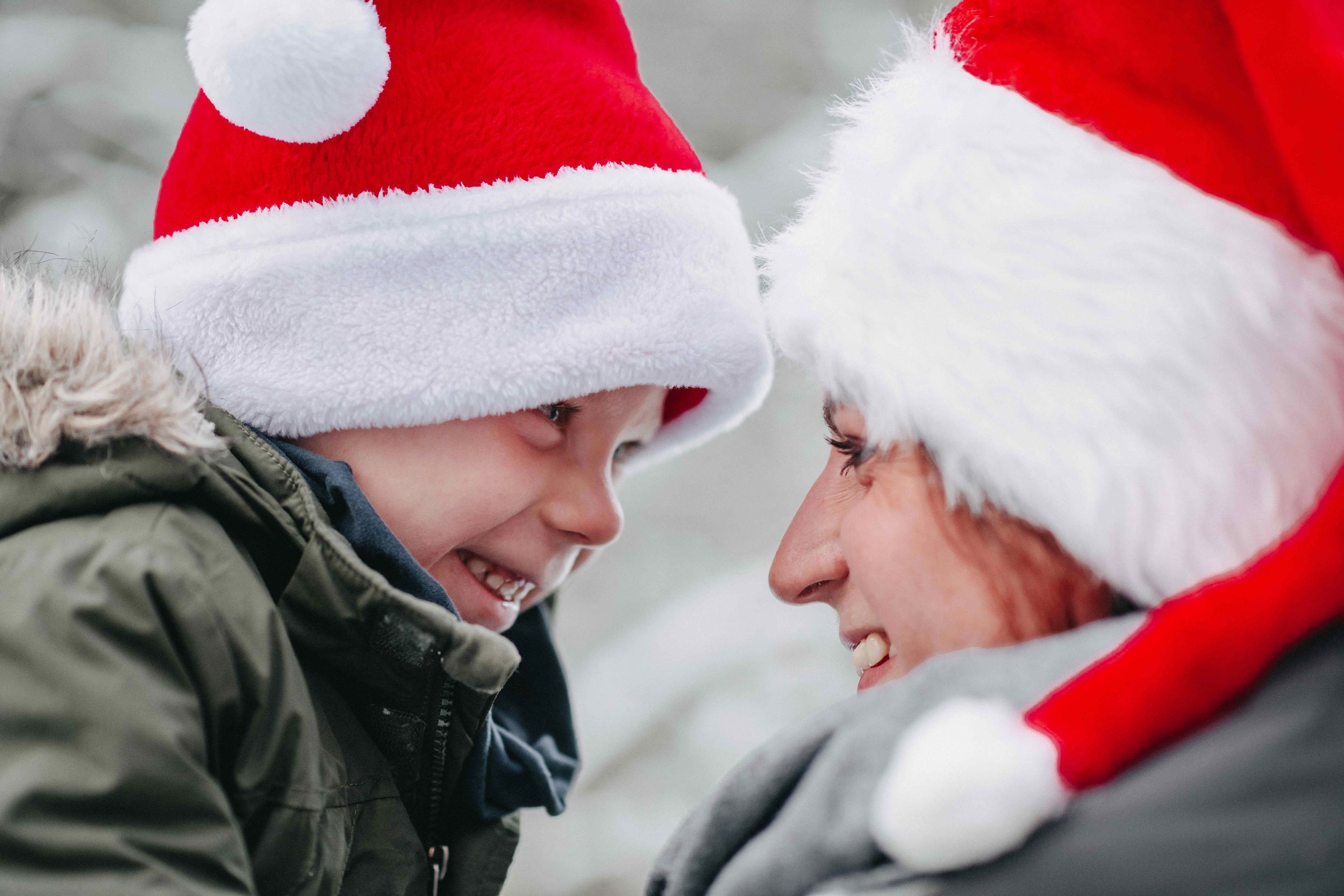 Ein Kind und seine Mama tragen rote Weihnachtsmützen und lächeln sich an.