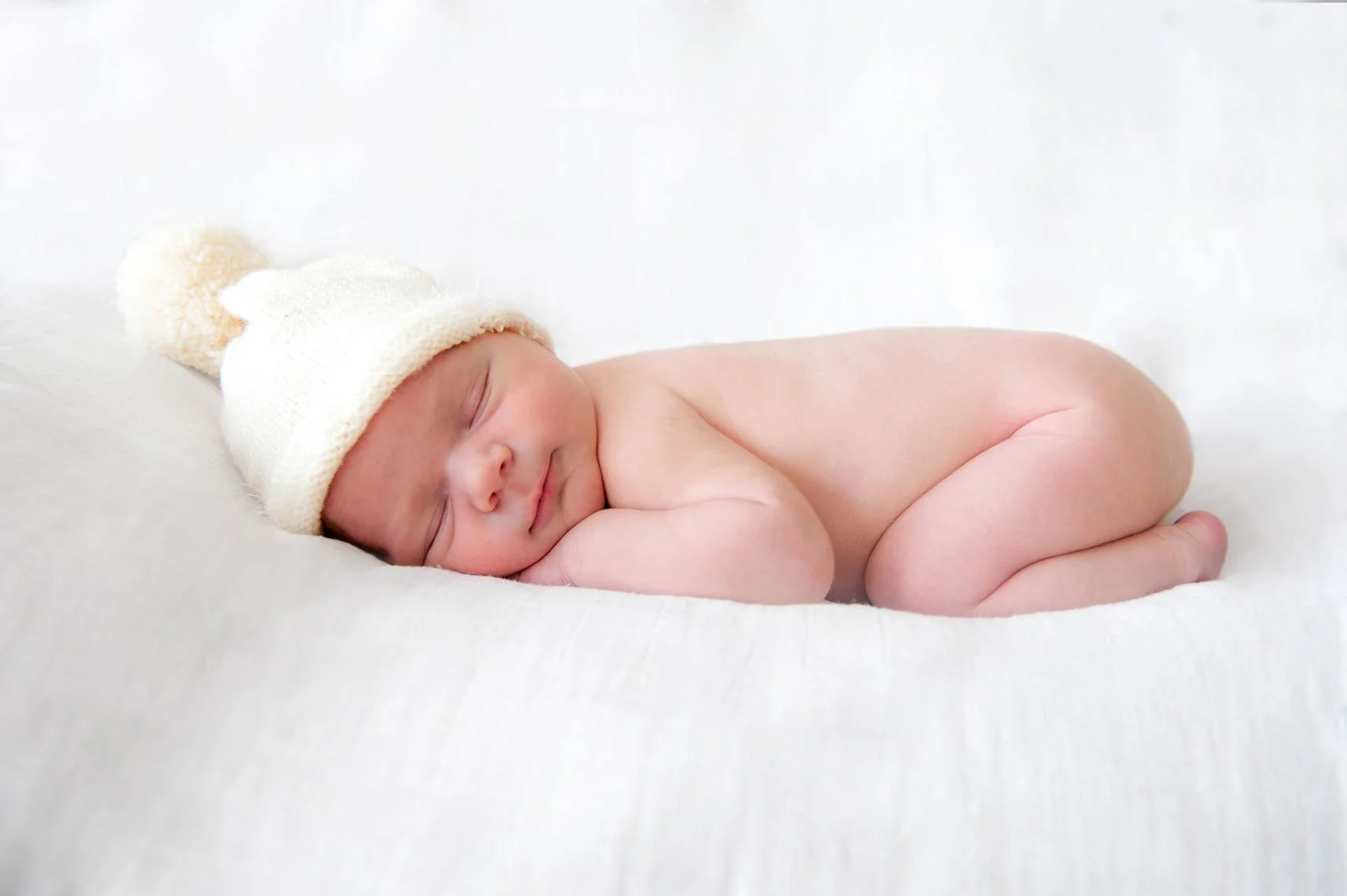 Baby nackt auf weisser Decke mit weisser Strickmütze.