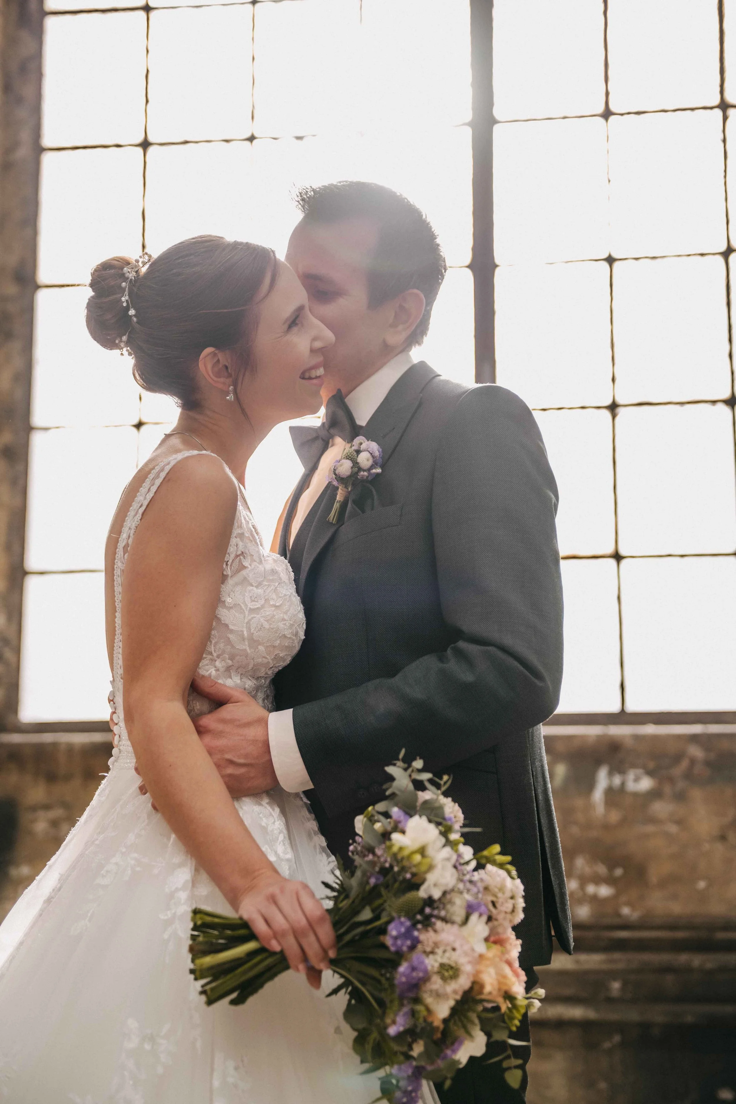 Hochzeitsfoto vom Brautpaar, das sich in einem Industrial-Style-Gebäude küsst, mit grossen Fenster im Hintergrund.