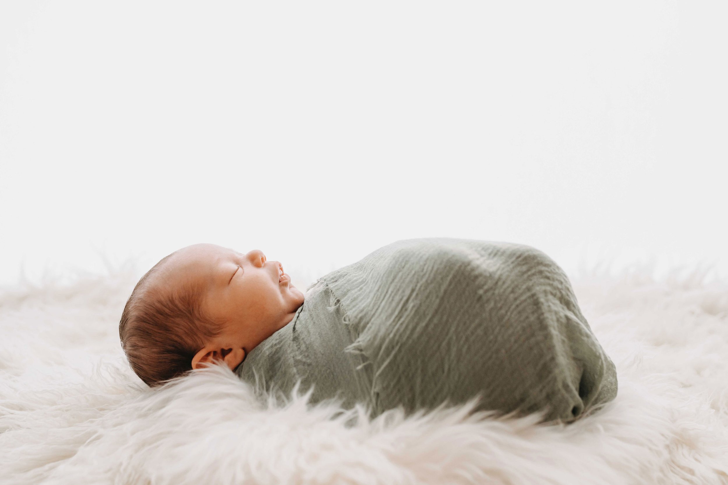 Schlafendes Baby in grünem Tuch eingewickelt auf einer flauschigen, weißen Decke liegend.