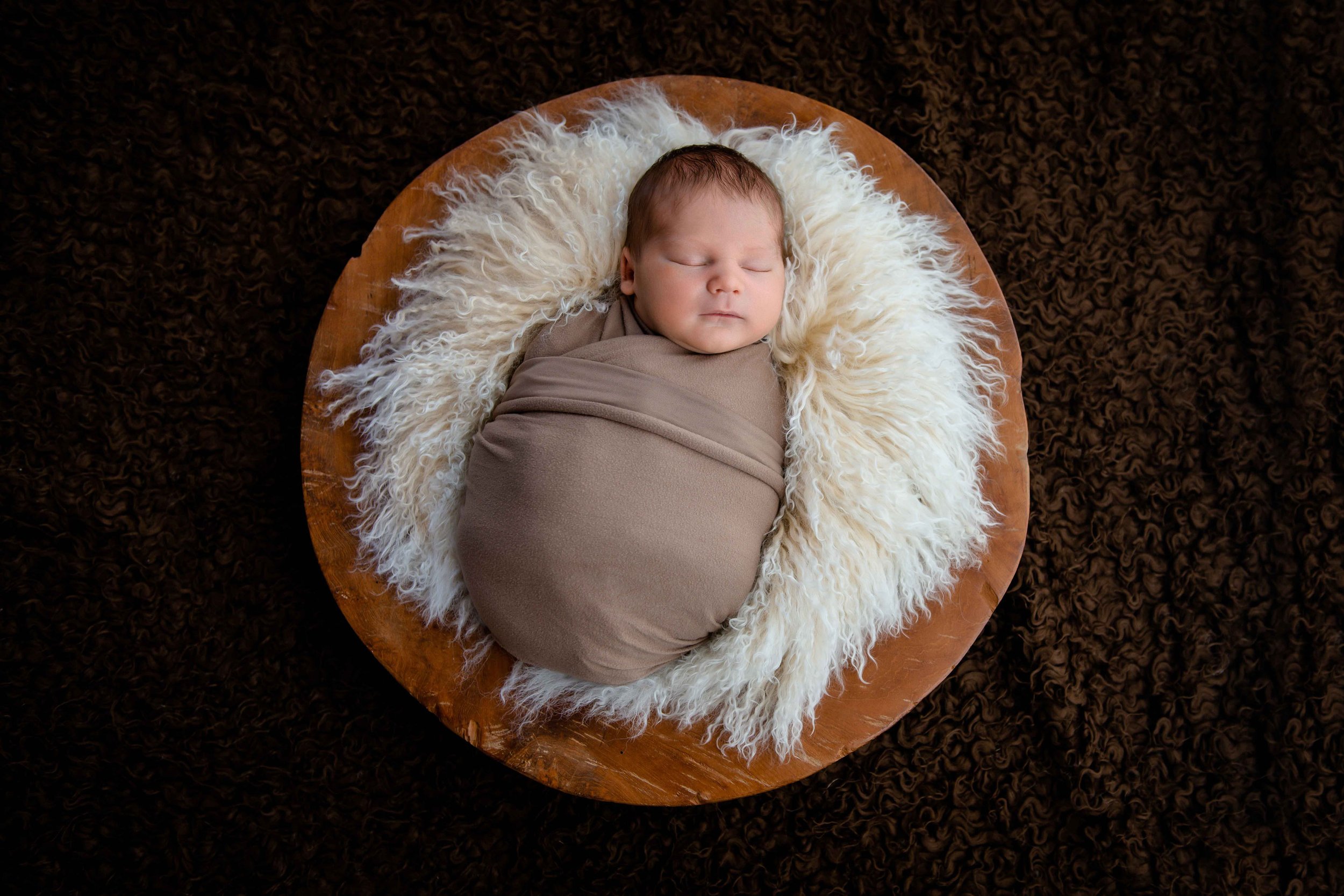 Schlafendes Baby in Decke eingewickelt auf flauschigem Fellkissen in Holzschale.