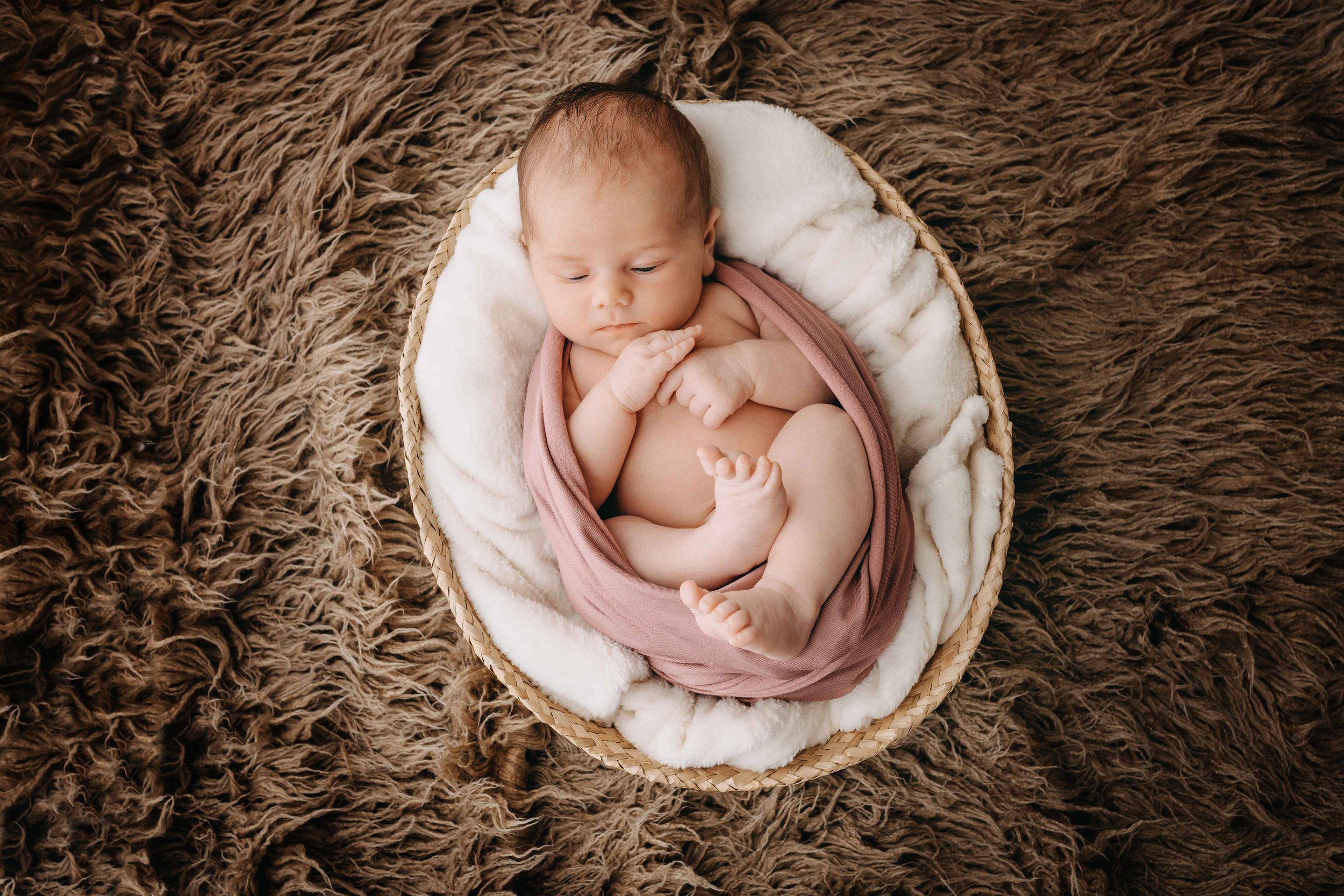 Neugeborenes Baby in einem Korb mit rosa Tuch auf braunem, flauschigem Teppich.