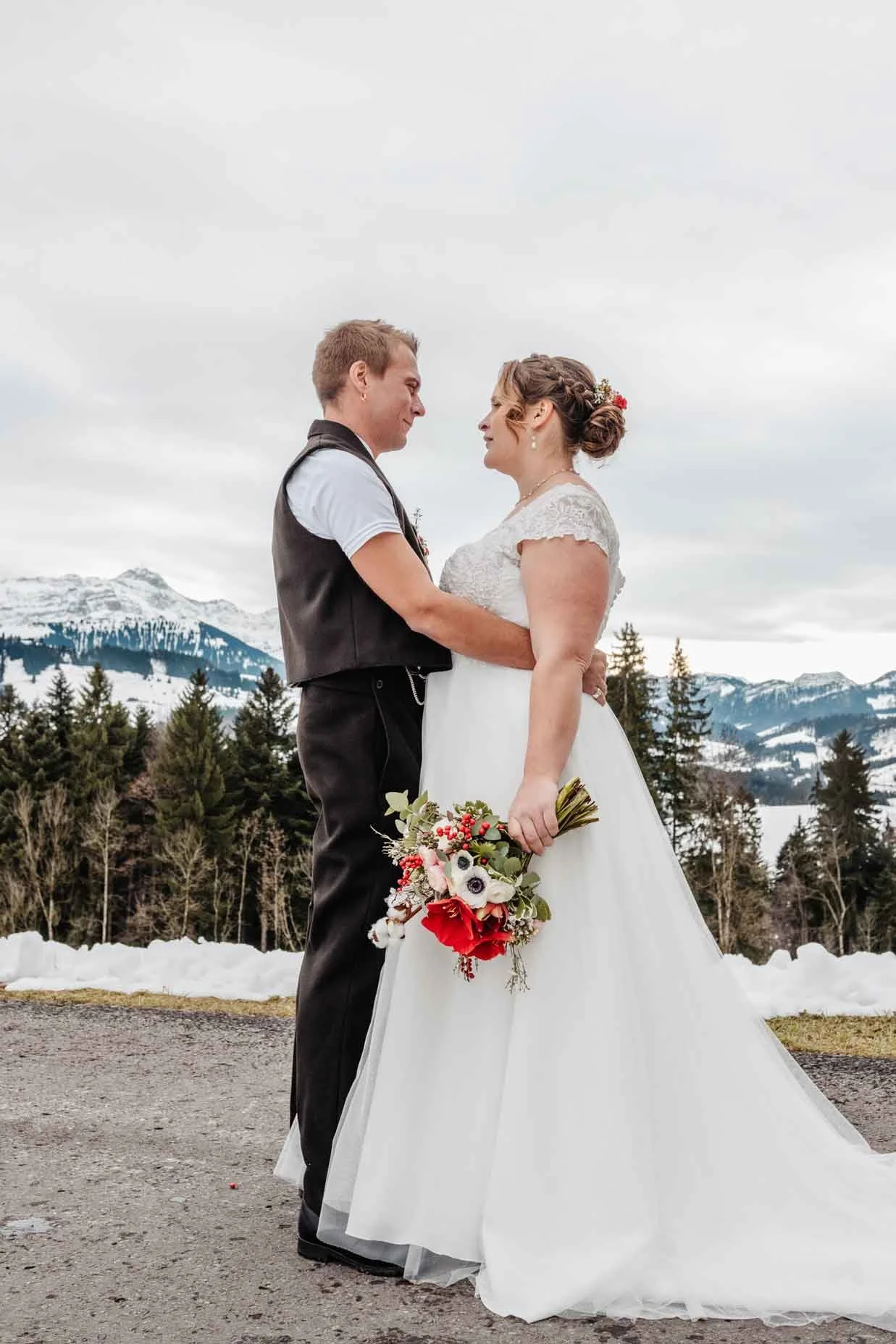 Brautpaar mit Bergen und Tannen im Hintergrund, bei Schneelandschaft. Die Braut trägt ein weisses Kleid, der Bräutigam trägt eine Bauerntracht.