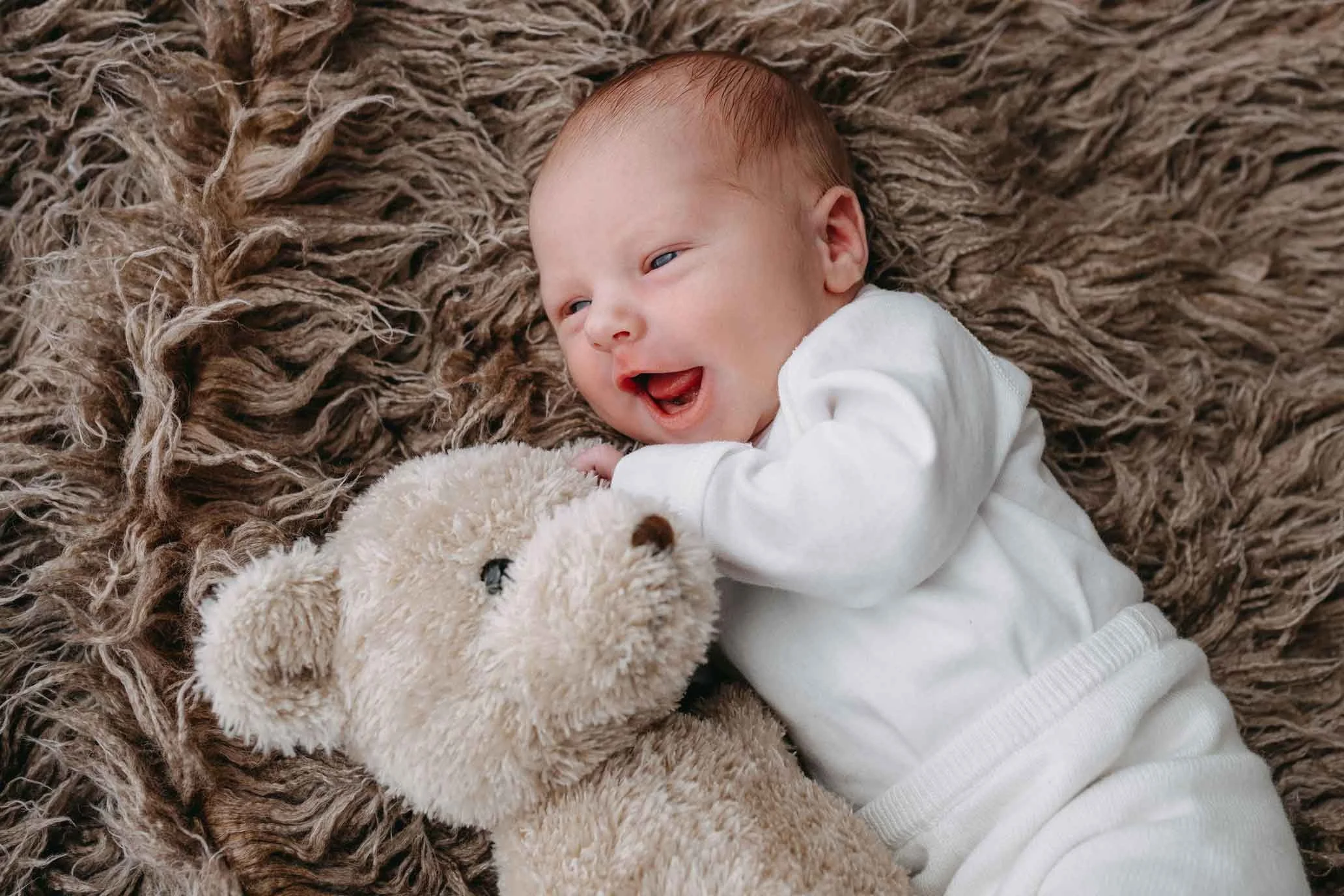 Lachendes Baby mit weißem Outfit liegt auf einem braunen, flauschigen Untergrund und kuschelt mit einem Kuscheltier