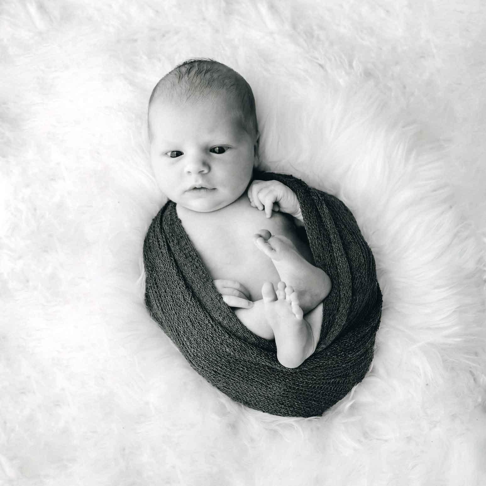 Schwarz-Weiss-Foto eines neugeborenen Babys, das auf einem weissen, pelzigen Hintergrund liegt, eng eingewickelt in eine dunkle Decke.