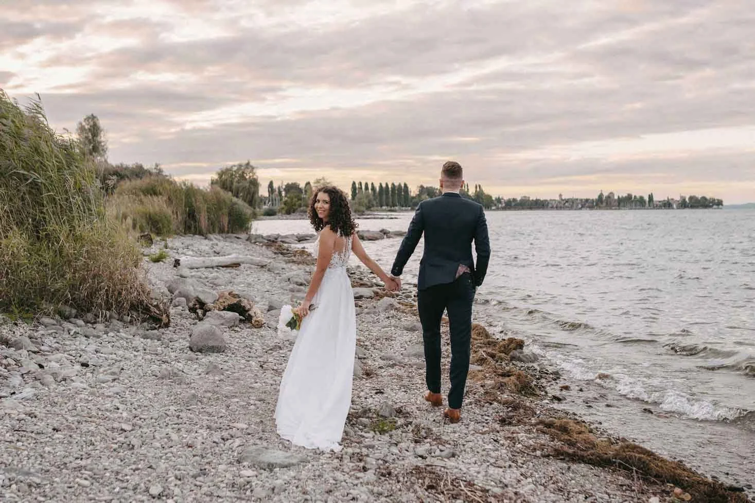 Paar in Hochzeitskleidung am Strand, die Hand halten, mit dem Meer und einem bewölkten Himmel im Hintergrund.