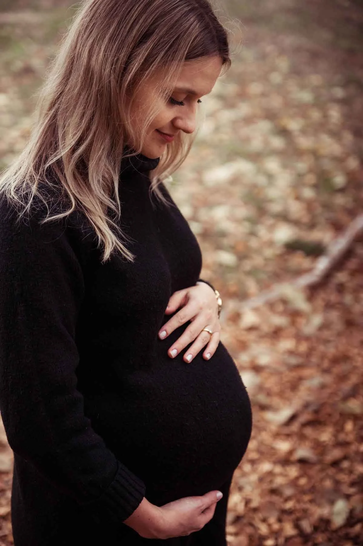 Schwangere Frau in einem schwarzen Pullover steht im Wald, hält ihre pralle Babykugel mit beiden Händen.