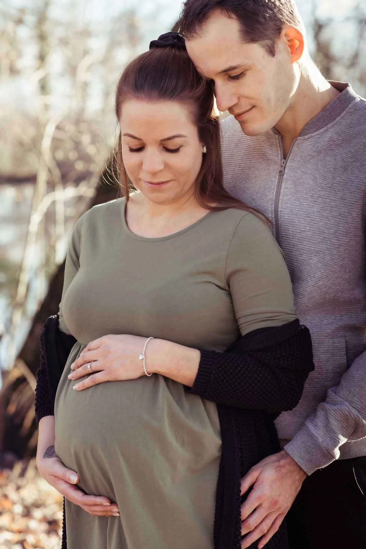 Ein schwangertes Paar steht im Freien im Sonnenlicht und hält ihren Babybauch, während der Mann sie umarmt und an den Bauch legt.
