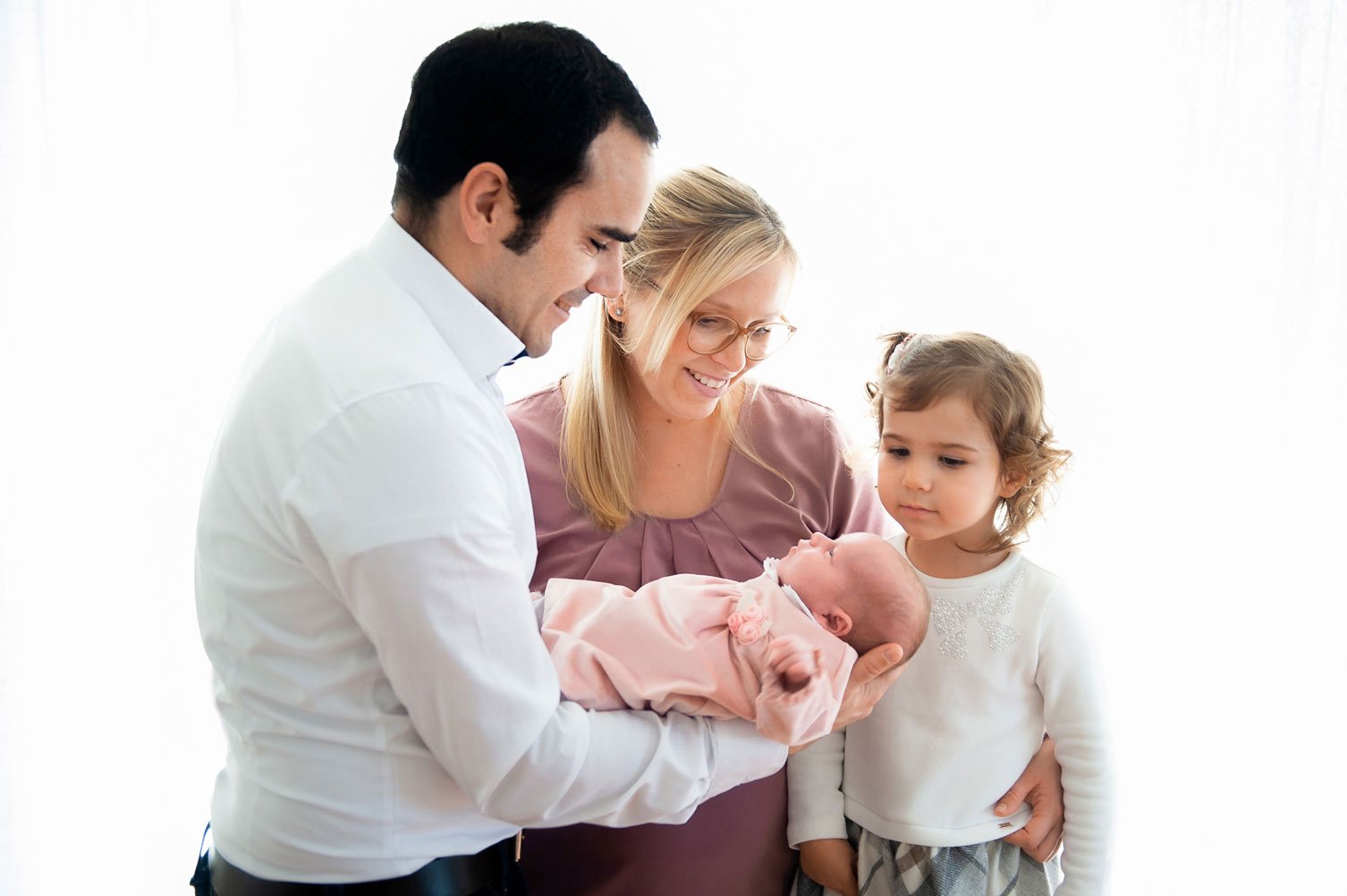 Lächelnde Familie mit Neugeborenem, Eltern und kleinem Mädchen, das in einem Raum zusammensteht.