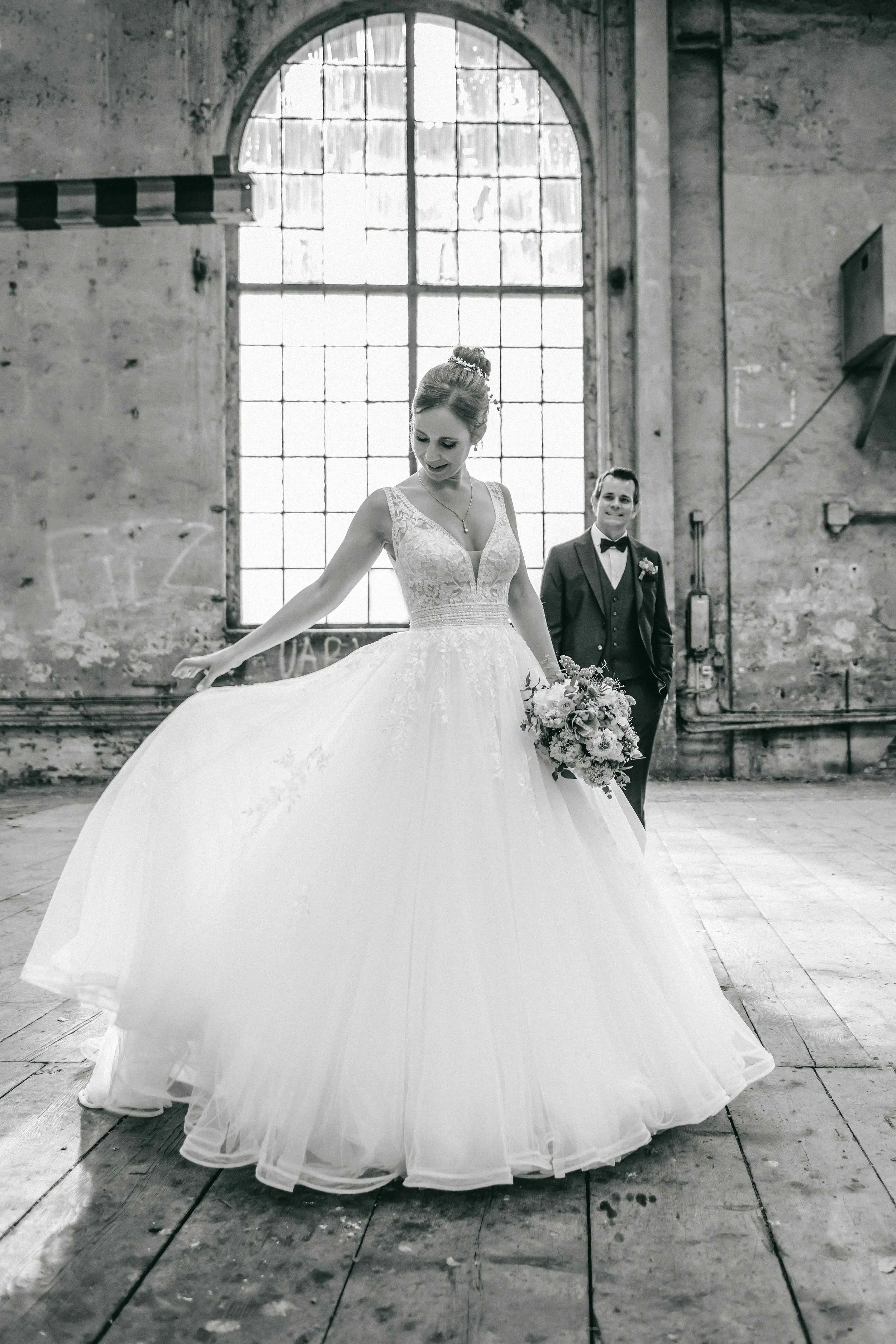Braut im Hochzeitskleid hält den Saum ihres weiten, weissen Kleides vor einem grossen Fenster in einer alten, rohen Industriehalle, während der Bräutigam im Anzug im Hintergrund lächelt.