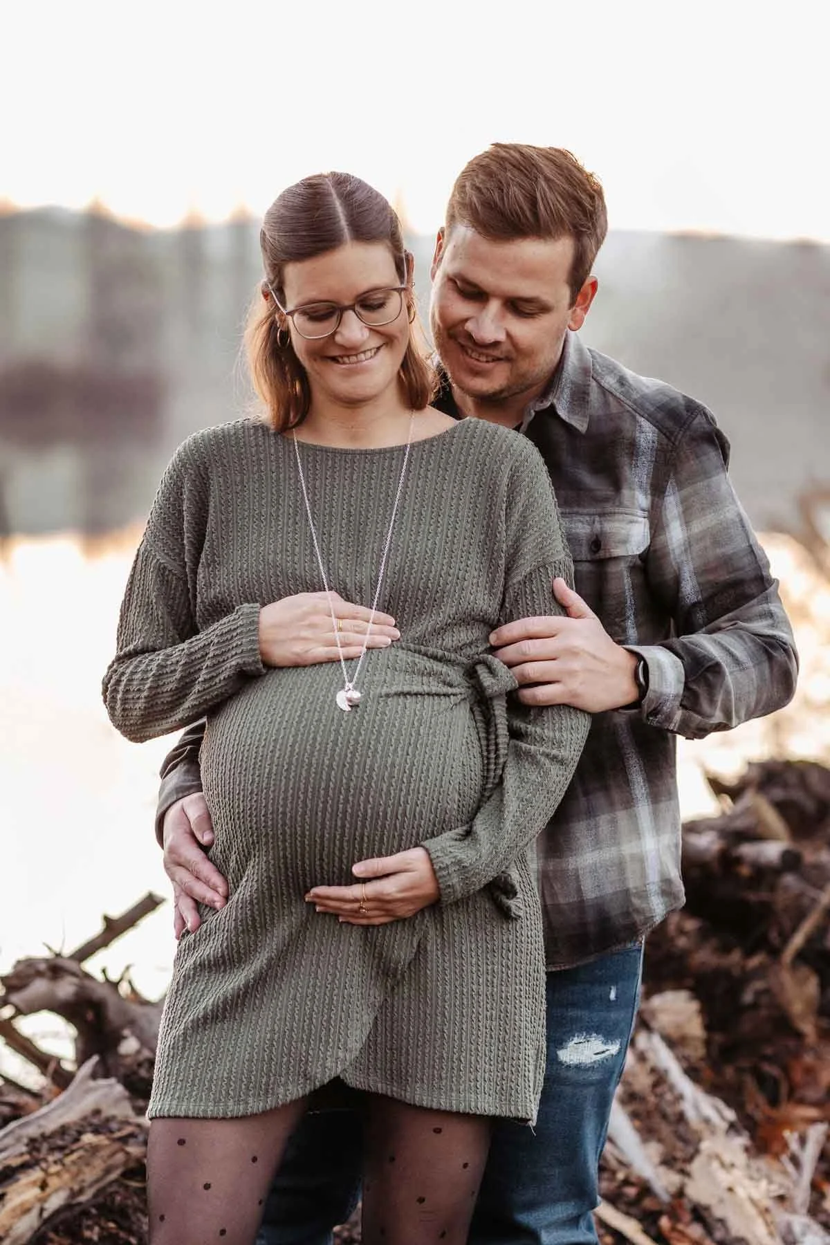 Ein Paar freut sich auf die Geburt ihres Babys, die Frau ist schwanger, sie stehen am Wasser bei Sonnenuntergang.