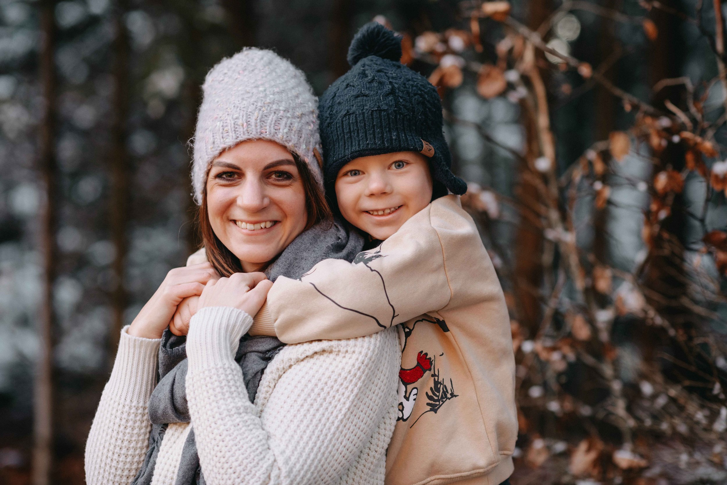 Mama und Kind im Winterwald, beide tragen Mützen und lächeln.