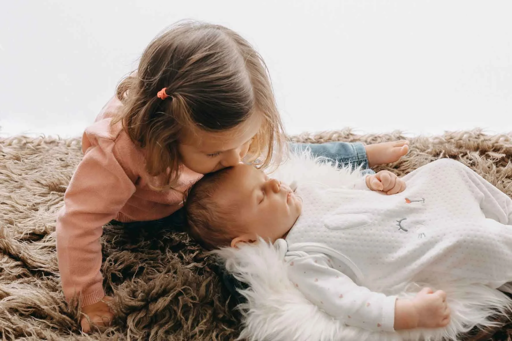 Ein kleines Mädchen küsst ein schlafendes Baby auf die Stirn, beide liegen auf einem flauschigen Teppich.