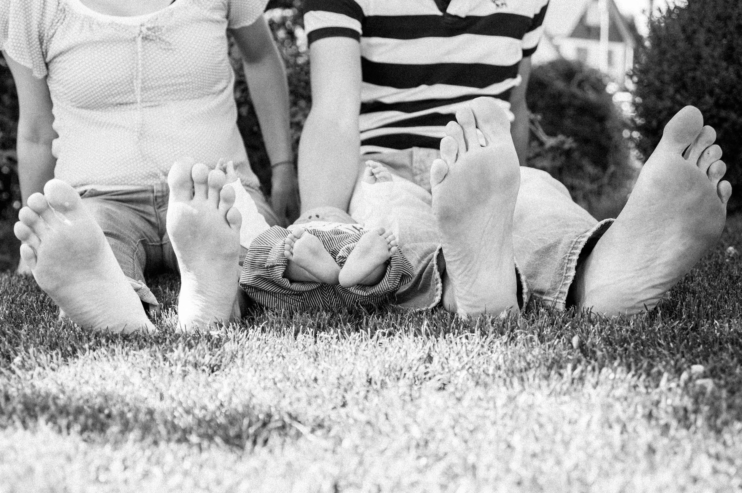 Schwarzweiss-Foto von drei Personen, die auf Gras sitzen. Die Eltern sind links und rechts mit ihren blossen Füssen ausgestreckt, in der Mitte ein Baby mit kleinen Füssen.