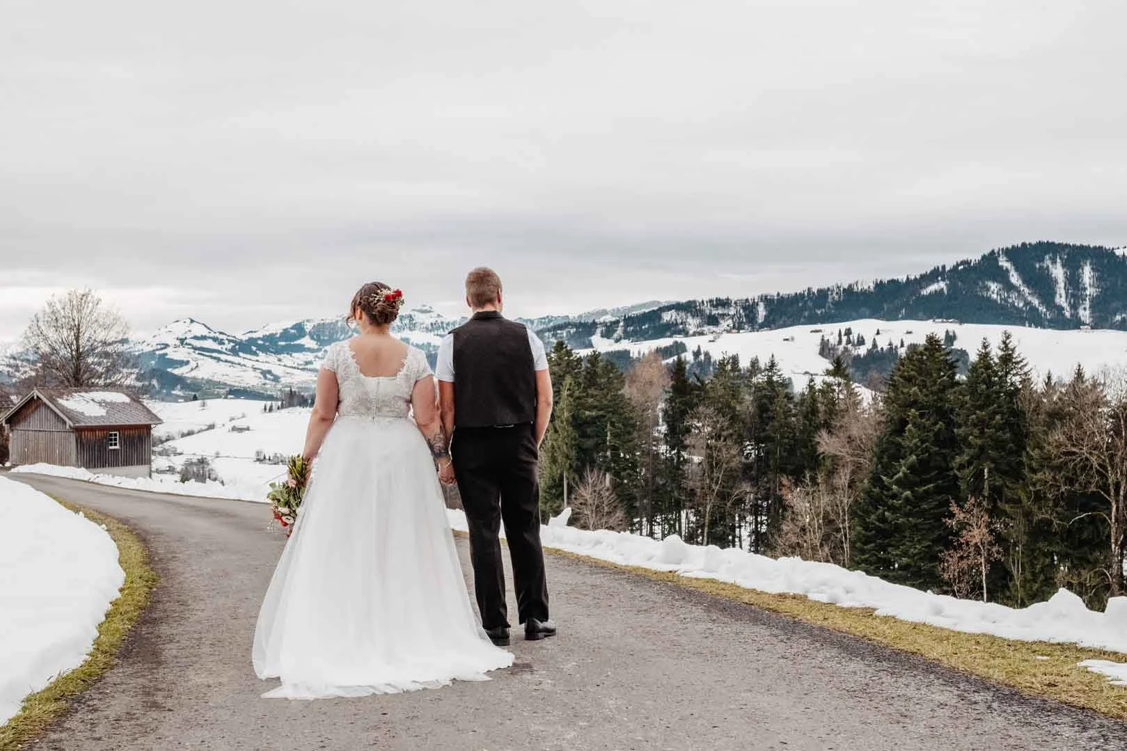 Brautpaar in Hochzeitskleidung geht Hand in Hand auf einer Landstraße in einer verschneiten bergigen Landschaft.
