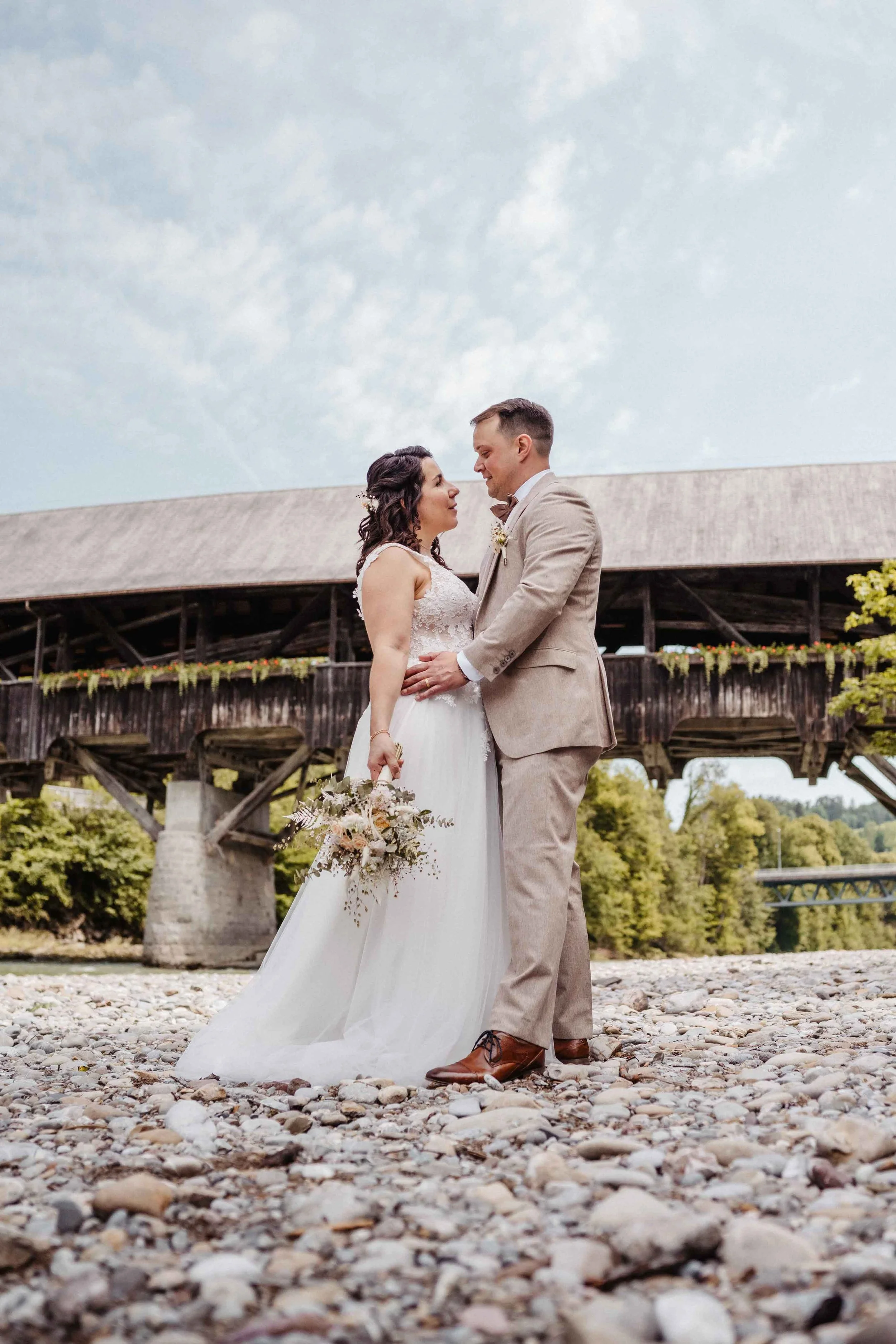 Ein Brautpaar steht sich am Fluss, die Braut trägt ein weißes Hochzeitskleid und hält einen Blumenstrauss, der Bräutigam trägt einen beige Anzug. Im Hintergrund ist eine alte Holzbrücke und der Himmel ist leicht bewölkt.