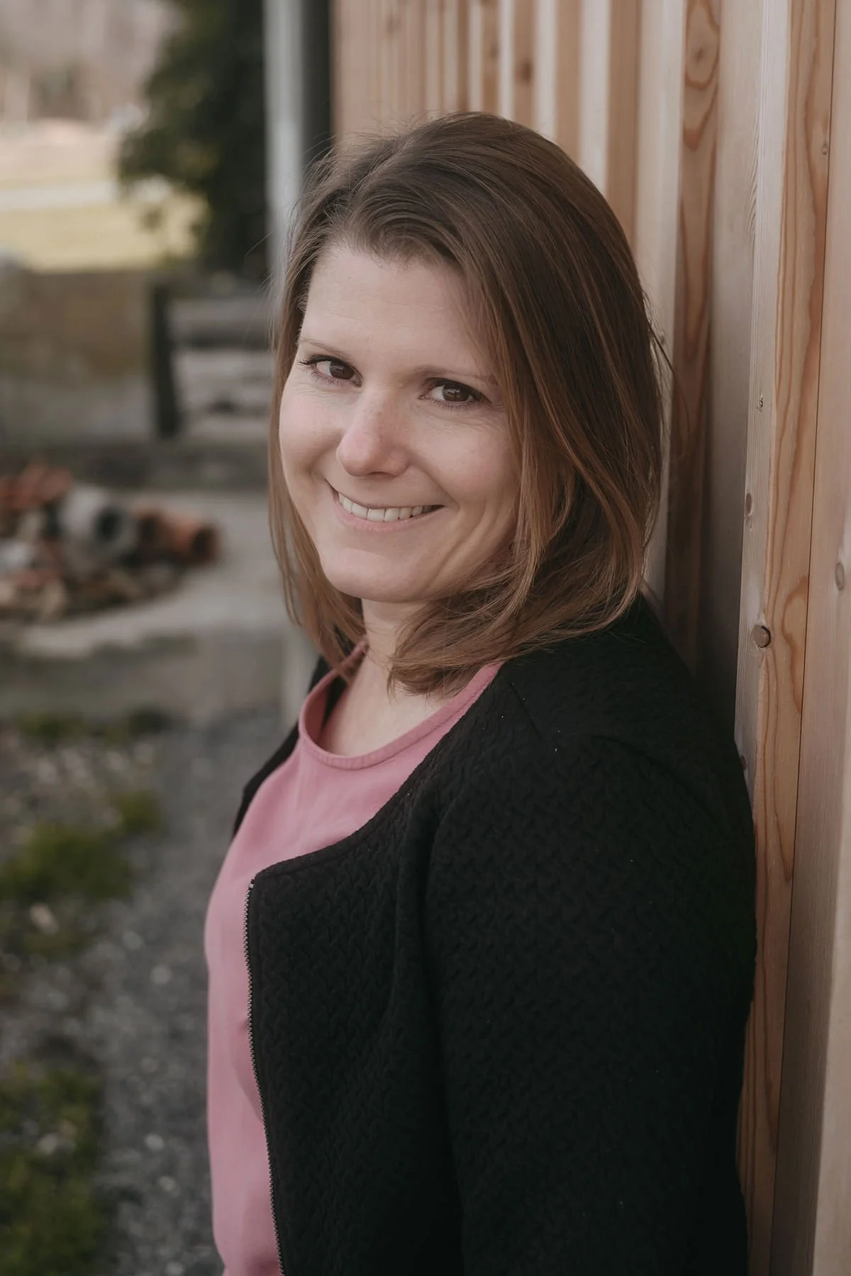 Eine lachende Frau mit braunen Haaren, trägt ein schwarzes Oberteil und eine pinkfarbene Bluse. Sie steht draußen neben einem Holzgebäude.