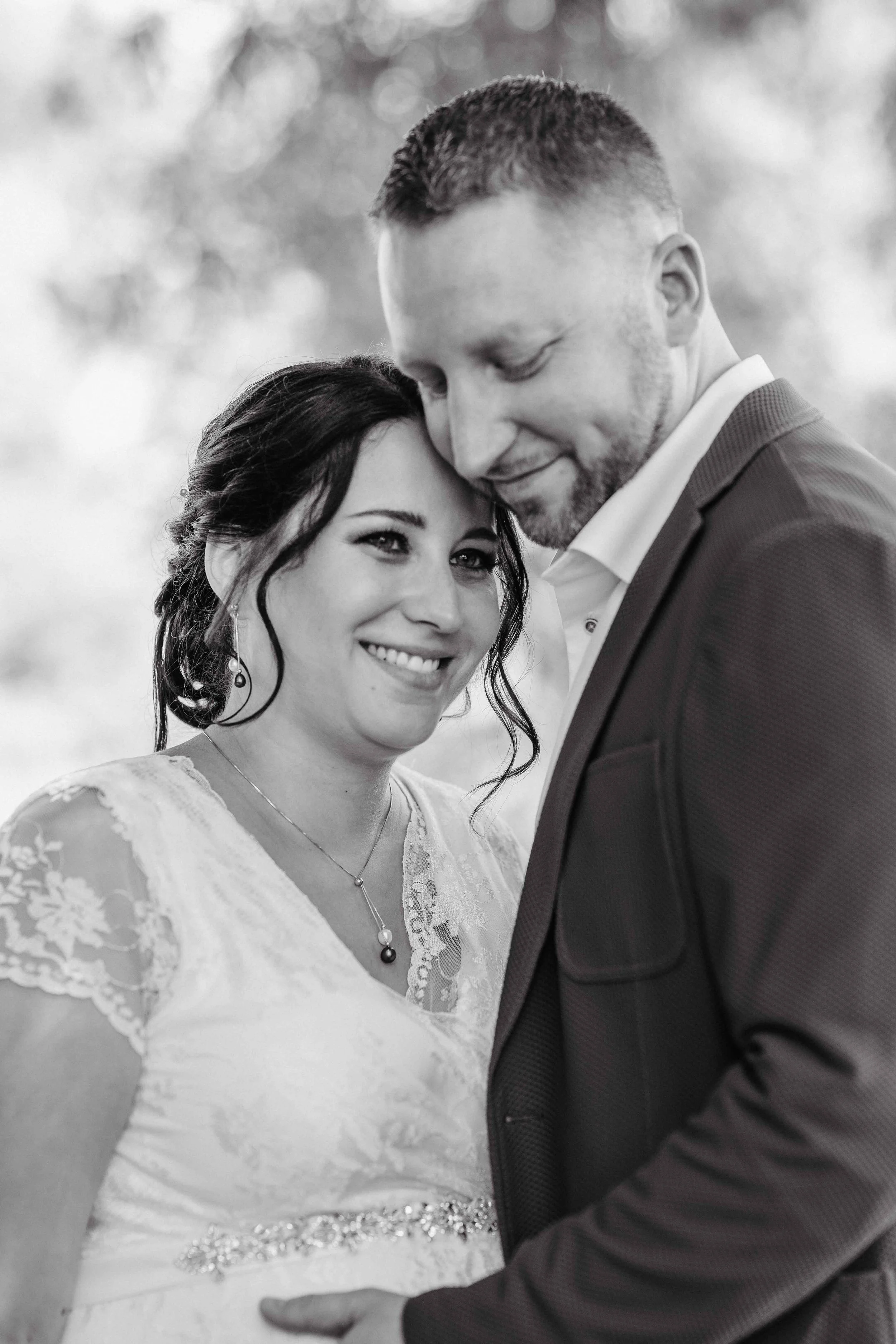 Brautpaar mit weissen Kleid mit Schmuck und Anzug, umarmt und lächelnd, schwarz-weiss Foto.