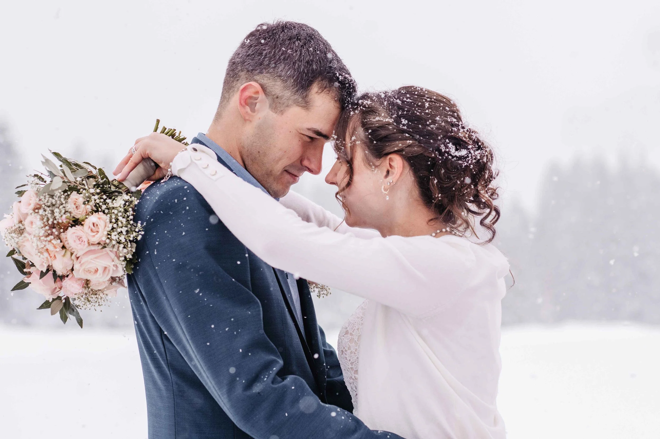 Ein Brautpaar in Schneeumgebung, umarmt und mit geschlossenen Augen, die Braut hält einen Brautstrauss aus rosa und weißen Blumen