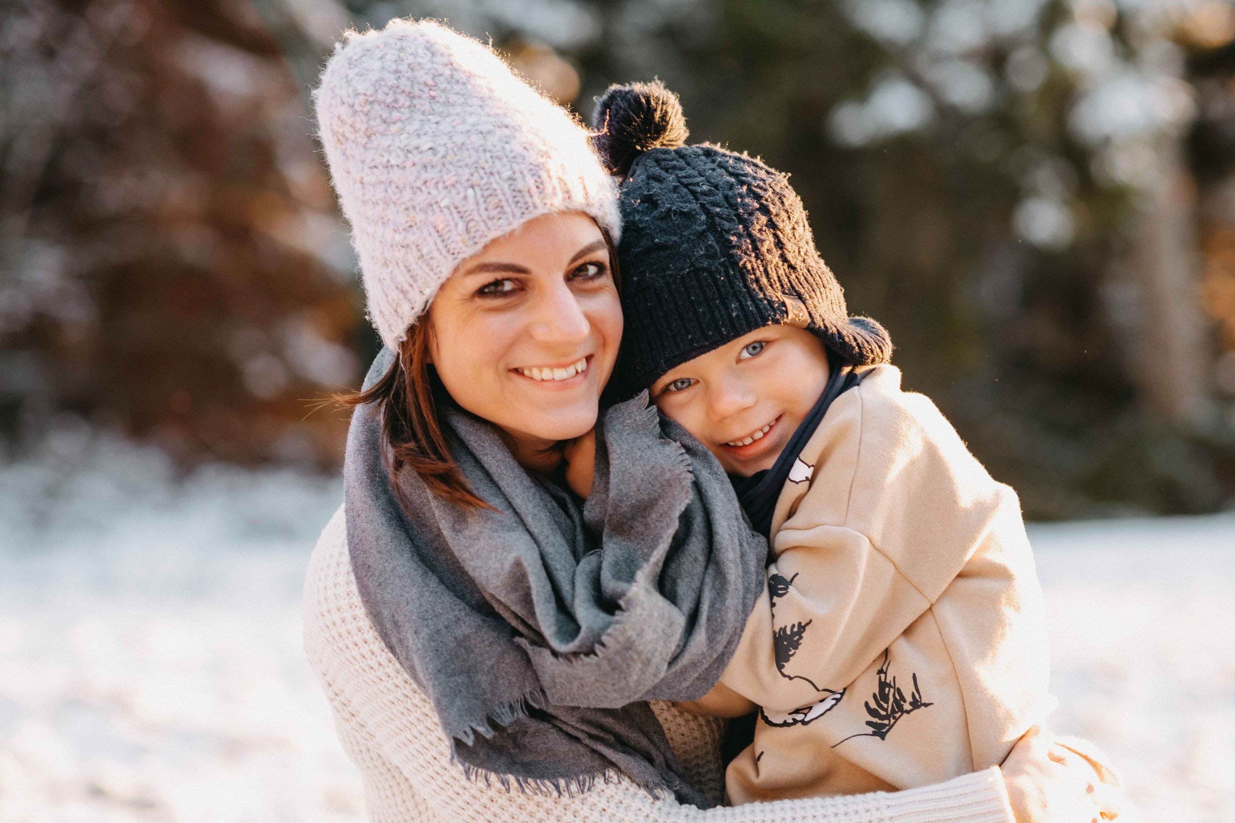 Eine Mama und ihr Kind in Winterkleidung, beide tragen gestrickte Mützen und lächeln. Sie sind draussen in einer schneebedeckten Umgebung.