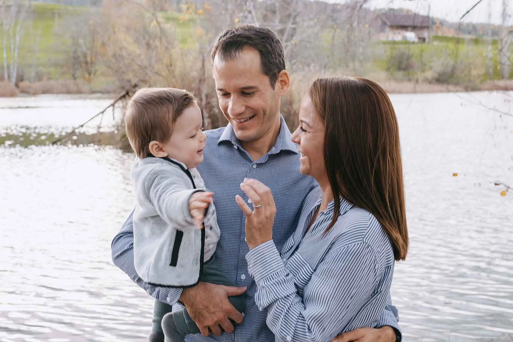 Familie steht am Fluss, lächeln und lachen, während ein kleiner Junge in den Armen seiner Mutter gehalten wird, im Hintergrund Bäume und ein Haus, herbstliche Stimmung