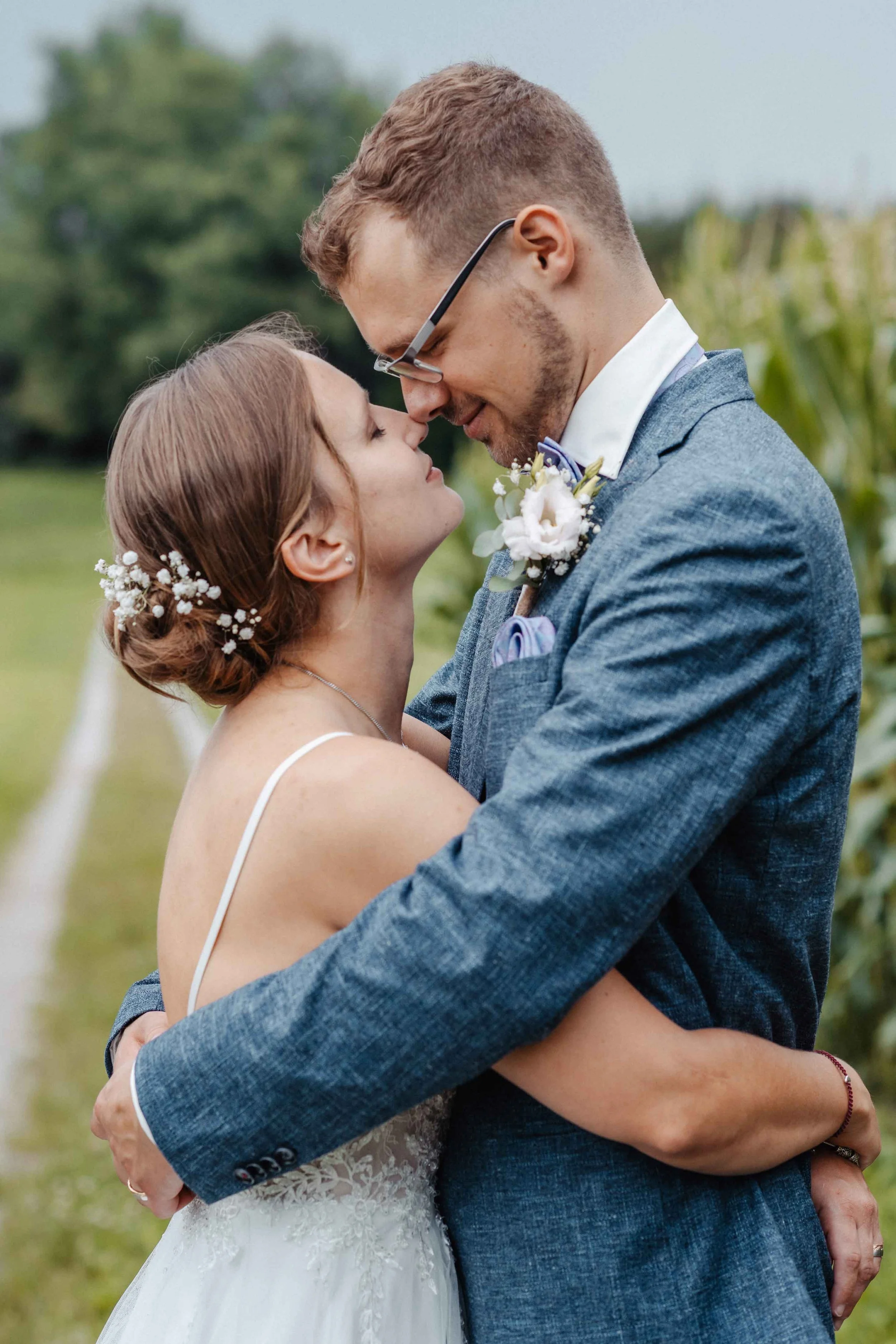 Brautpaar eng umarmt. Im Hintergrund sind grüne Bäume und ein bewölkter Himmel sichtbar.